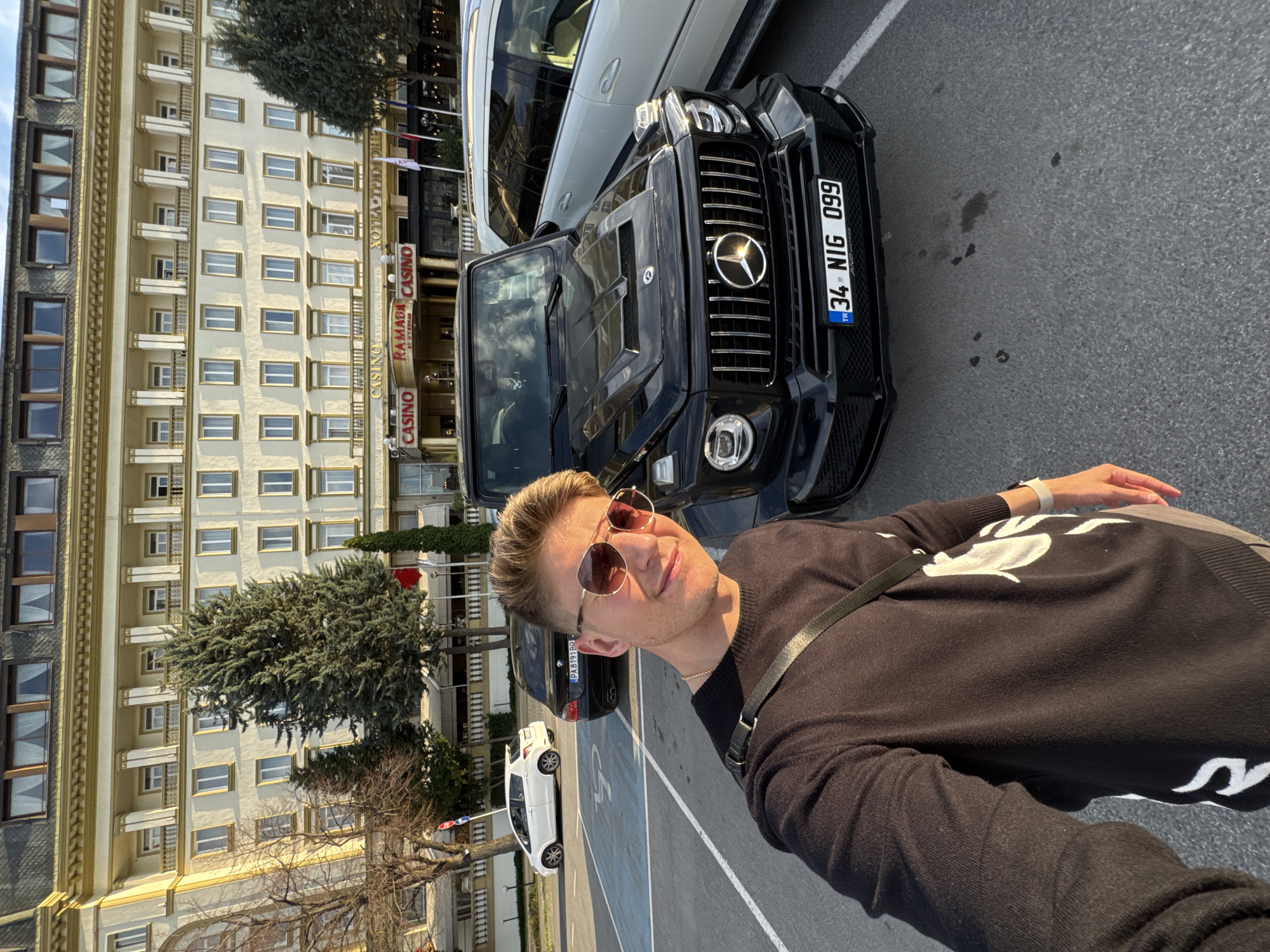 A man wearing sunglasses and a dark sweater stands on a gray surface with white painted lines, likely a parking area. He is taking a selfie with a black luxury SUV behind him, revealing a license plate that reads "34 NIG 099." In the background is the Ramada Plovdiv Trimontium Casino and hotel. This suggests the location is Plovdiv, Bulgaria. The sky is visible, and there are trees nearby. Other cars are parked further in the background.