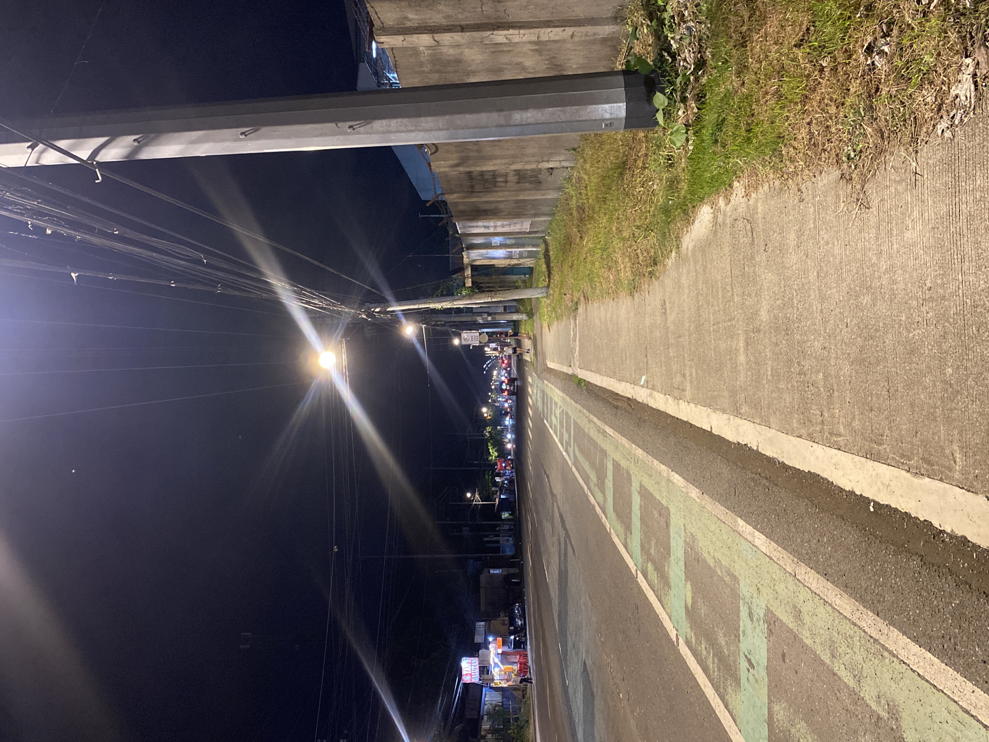 The image captures a street scene at night in Davao City, Philippines. The perspective is from a sidewalk, looking down a paved road that has a dedicated lane marked with green rectangles. Several streetlights illuminate the scene, casting long streaks of light due to lens flare. On the left side of the road, there are small food stalls with people, and further down, traffic is visible. On the right side of the sidewalk, a tall utility pole stands next to a concrete wall. There is also overgrown grass and vegetation along the edge of the sidewalk and wall. The sky is dark, indicating nighttime.