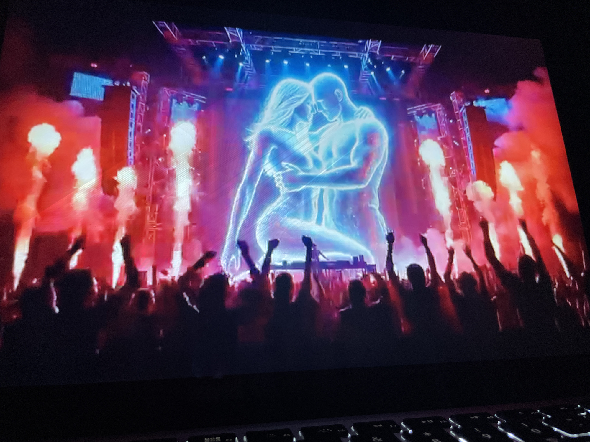 A luminous, ethereal image of a man and woman embracing is projected onto a large screen at a concert or festival. The couple is rendered in bright blue light, appearing almost as spirits. They are positioned centrally on the screen, with the woman on the left and the man on the right, their heads tilted towards each other. Behind them, a vibrant stage setup is visible, with rows of lights and trussing. Red and pink smoke or lighting effects billow outwards from the sides of the screen, creating a dramatic atmosphere.
In the foreground, silhouetted against the bright screen, is a large crowd of people. Many in the crowd have their arms raised in the air, suggesting excitement and engagement with the performance. The overall impression is one of a lively and energetic event.
The question of location in Kason, Nigeria, cannot be answered from the image alone. The image depicts a screen display and a crowd, but there are no discernible landmarks, signs, or other contextual clues that would indicate a specific location within Kason or any other city.