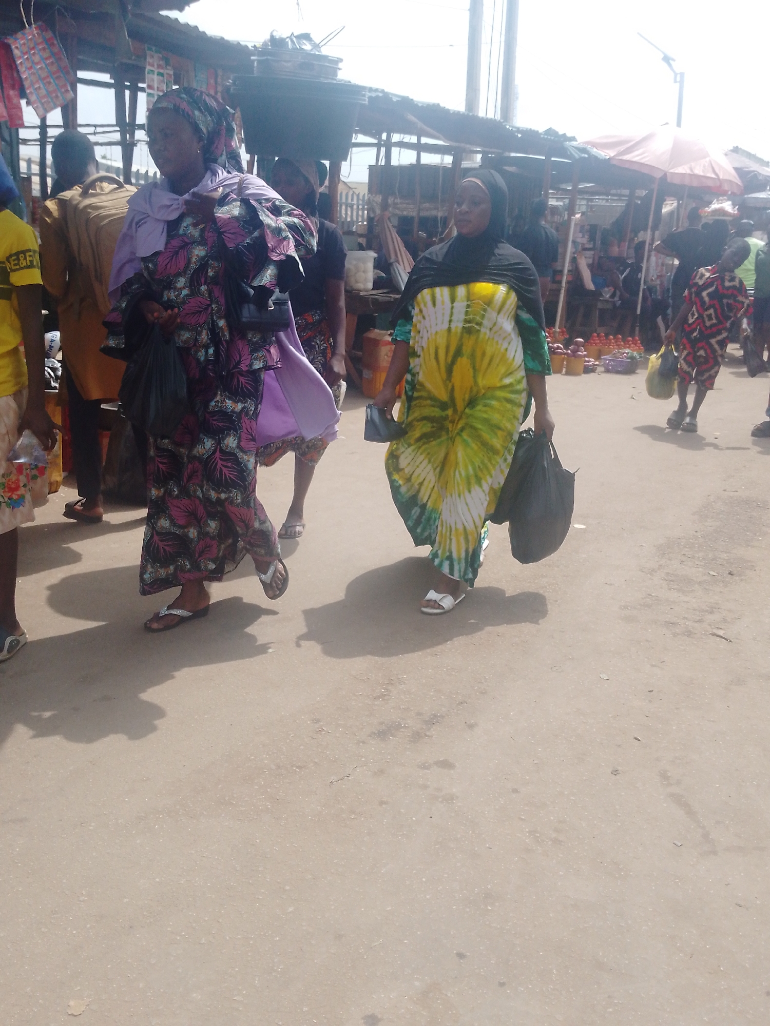 In an open-air market in an unspecified city, two women dominate the foreground. The woman on the left is dressed in a long patterned dress with shades of black, purple, and pink, topped with a lilac scarf. She is carrying a large bundle of material and a black purse. The woman on the right wears a long yellow and white tie-dye dress with a black hijab and carries two black plastic shopping bags. Both women are wearing flip-flops. The background is filled with other people, market stalls covered by makeshift roofs, and a red umbrella. The ground is a light-colored, paved surface, with shadows of the figures cast towards the bottom of the frame.