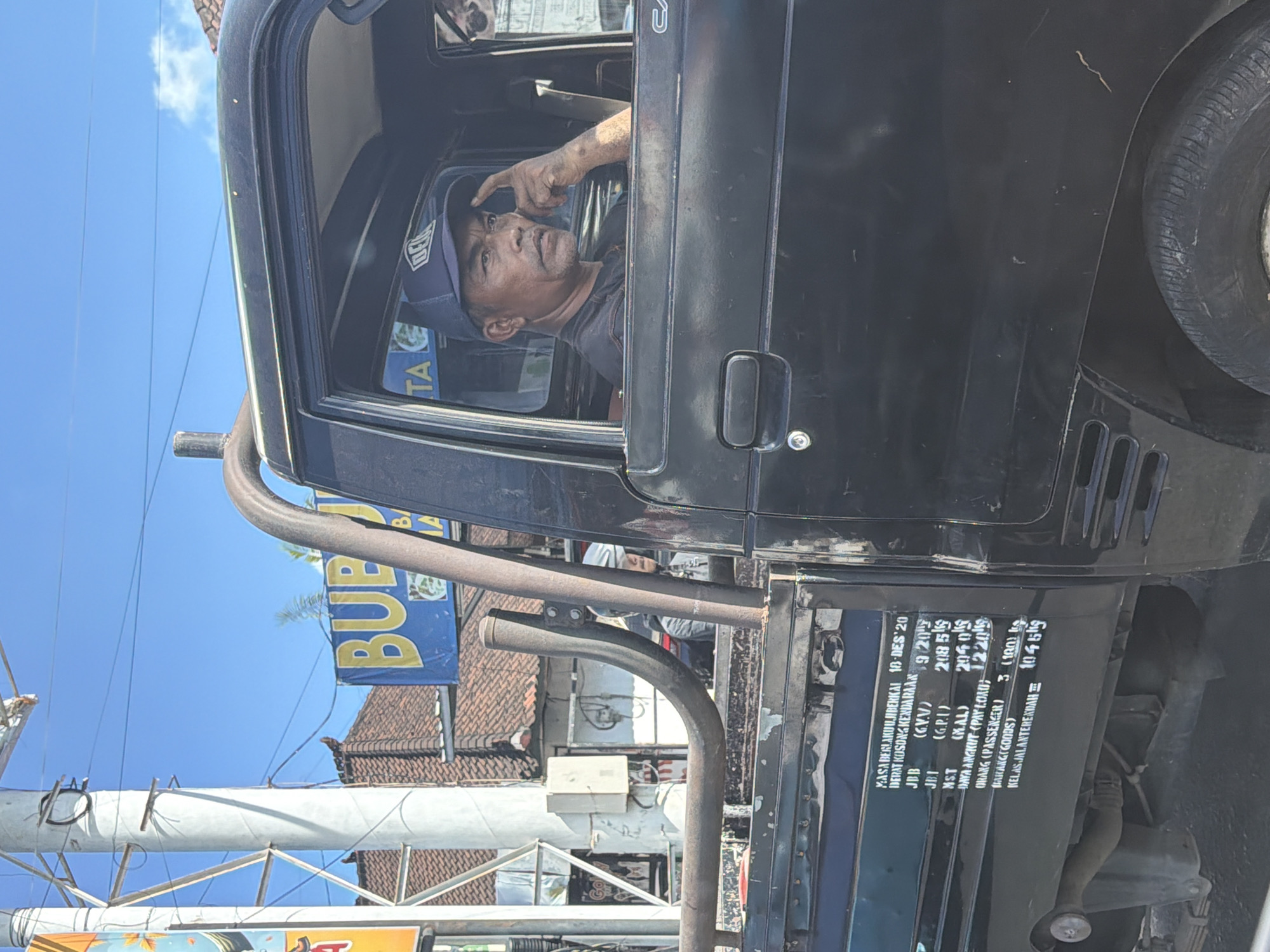 The image captures a close-up of a man inside a dark-colored utility vehicle or pickup truck, set against a bright blue sky in North Kuta, Indonesia. The man, appearing middle-aged or older with a weathered face, wears a dark blue baseball cap and a dark t-shirt. He looks out the open driver's side window with a neutral expression, his right hand slightly raised with his index finger near his temple or cap brim.
The black vehicle features custom-fitted metal pipes or roll bars along its side, and a spare tire is visible towards the rear. A white sticker or plate affixed to the lower rear side of the truck displays various specifications in Indonesian. Visible text includes:
* "MASA BERLAKUUJIBERKAI 18-DES 20" (Validity period of roadworthiness test 18-Dec-20)
* "DERAT KOSONG KENDARAAN 920" (Empty vehicle weight 920 kg)
* "JBB (GVV) 2085 kg" (Gross Vehicle Weight)
* "JBI (G.P.1) 2040 kg" (Gross Permissible Weight)
* "MS.T (MAL) 2040 kg" (Max. Axle Load)
* "DAYA ANGKUT (PAYLOAD: 1220 kg" (Payload capacity)
* "ORANG (PASSENGER) 3 (100 kg)" (Number of passengers and total weight)
* "BARANG(GOODS) 1045 kg" (Goods weight)
* "KELAS JALANTERENDAH Ξ" (Lowest road class)
The background shows a clear blue sky with thin power lines strung across. Tiled rooftops of buildings are visible, along with a blue and yellow banner partially displaying "BUP", likely part of the word "BUBUR" (porridge). The strong sunlight indicates it is daytime, and the overall scene suggests a moment of rest or waiting in a bustling, semi-urban environment.