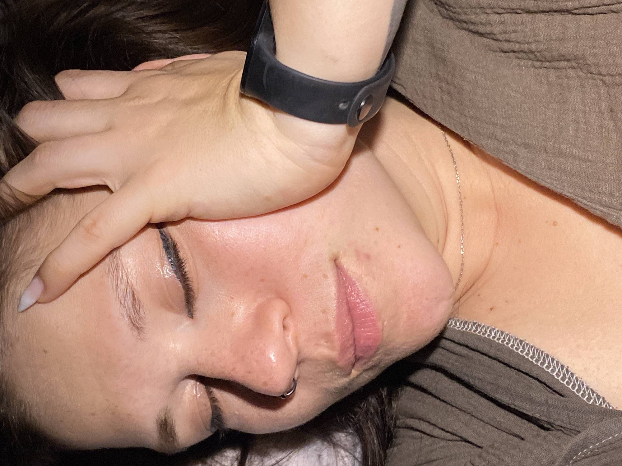 A close-up, head-and-shoulders shot focuses on the right side of a person's face, with their eyes closed. Their right hand is placed on their forehead, partially obscuring it, and their fingers are tangled in dark hair. A black watch is visible on their wrist. The person is wearing a brown, textured garment. A thin silver necklace is visible at the base of their neck. The background is dark and out of focus. The image does not contain any elements that would allow for identification of its location in Chaikovsky, Russia.