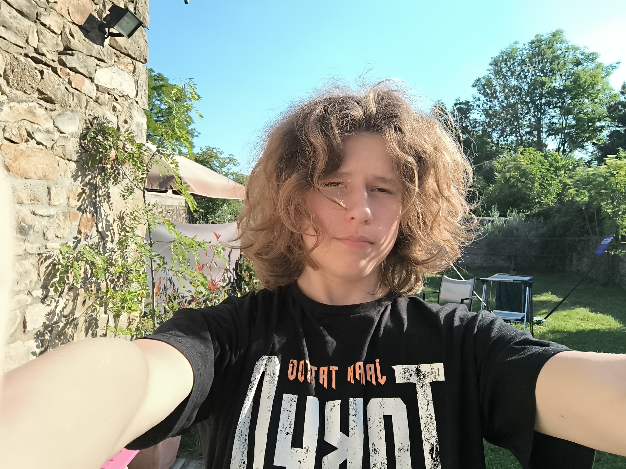 A young person with curly brown hair is taking a selfie outdoors. The person is wearing a black t-shirt with large, distressed white lettering and orange details. The background features a stone building on the left, with climbing plants. To the right of the person is a grassy area with trees and what appear to be camping chairs and a table. The sky is blue. The location is potentially Pélussin, France given the query, but no specific elements in the image confirm this location directly. The focus is on the person and their immediate surroundings.