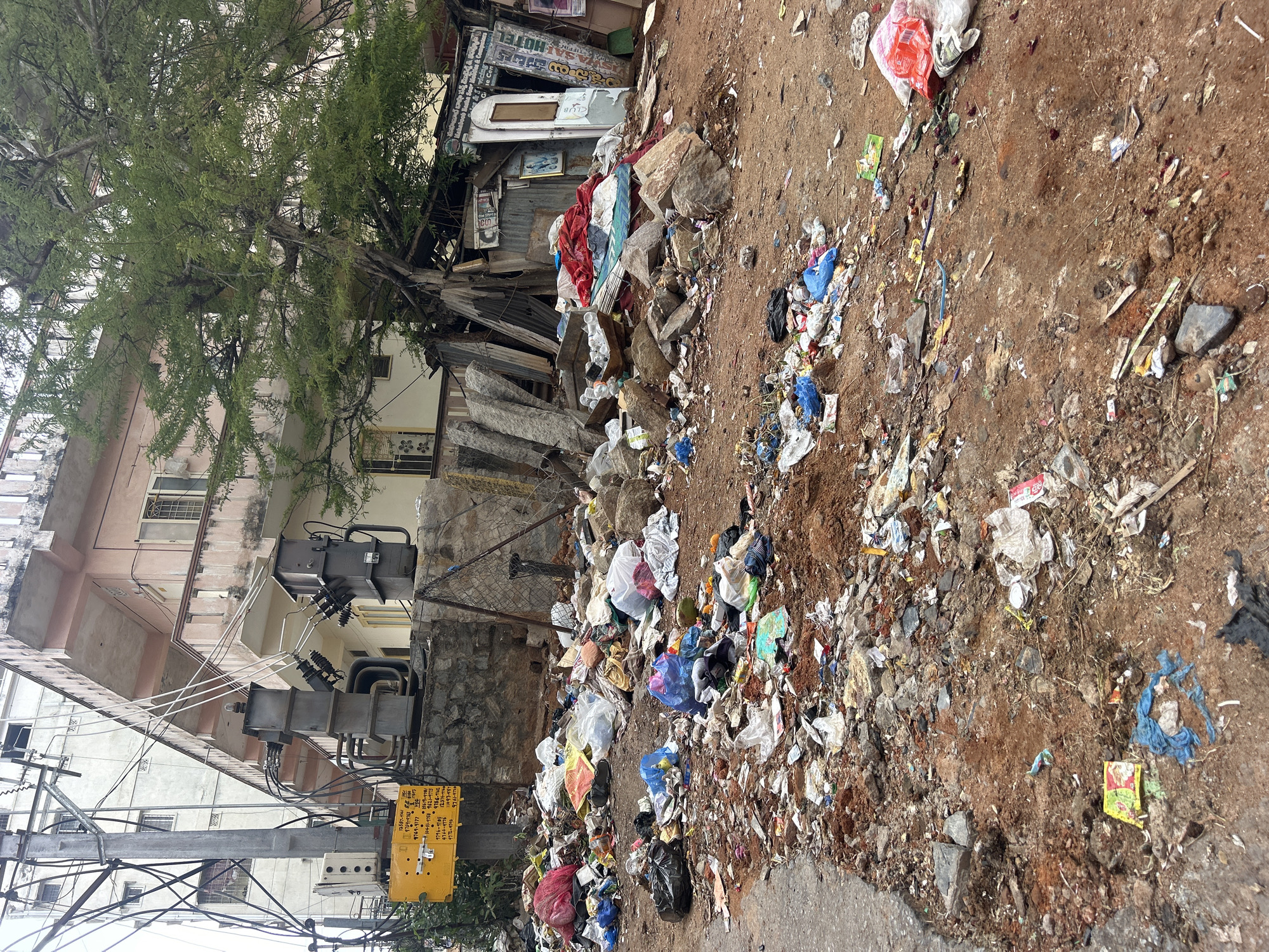 The image depicts a scene of significant urban decay and neglect in Puttaparthi, India. In the foreground and midground, a vast amount of refuse, including plastic bags, debris, household waste, and what appear to be discarded textiles, is piled haphazardly on a dirt and asphalt surface. The trash is spread across the ground, obscuring parts of the road and creating an unsanitary environment.
In the background, a multi-story building with a light pink facade and balconies stands alongside electrical infrastructure, including utility poles, transformers, and a dense network of wires. Adjacent to the building and integrated with the refuse is a ramshackle structure, possibly a makeshift stall or shelter, constructed from corrugated metal and wooden planks. This structure is also cluttered with debris and advertising signage in an Indian script. A large tree with green foliage is growing intertwined with the debris and the structure, its branches reaching towards the building. A yellow informational sign with text in an Indian script is mounted on a pole near the electrical equipment. The overall impression is one of significant environmental degradation and a lack of municipal sanitation in a densely populated urban area.