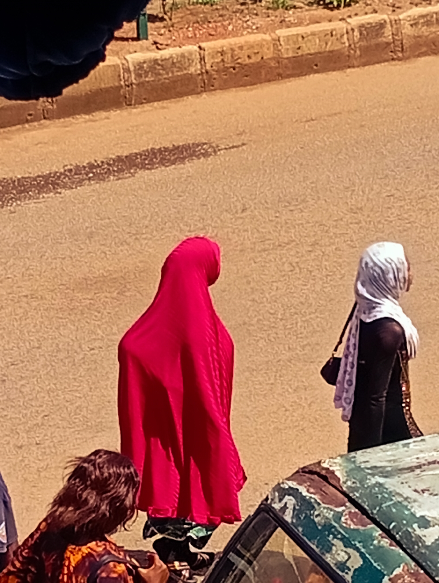 In Bukuru, Nigeria, two women are walking on a street. The woman on the left is wearing a bright pink garment that covers her head and body. The woman on the right is wearing a black top and a white hijab. She has a small dark purse with a patterned strap. A person with dark, wavy hair is visible in the foreground on the left, wearing a patterned top. In the lower right of the image, the roof of a car with peeling paint is visible. The street is paved with asphalt, and a concrete curb with a dirt shoulder is visible in the background.