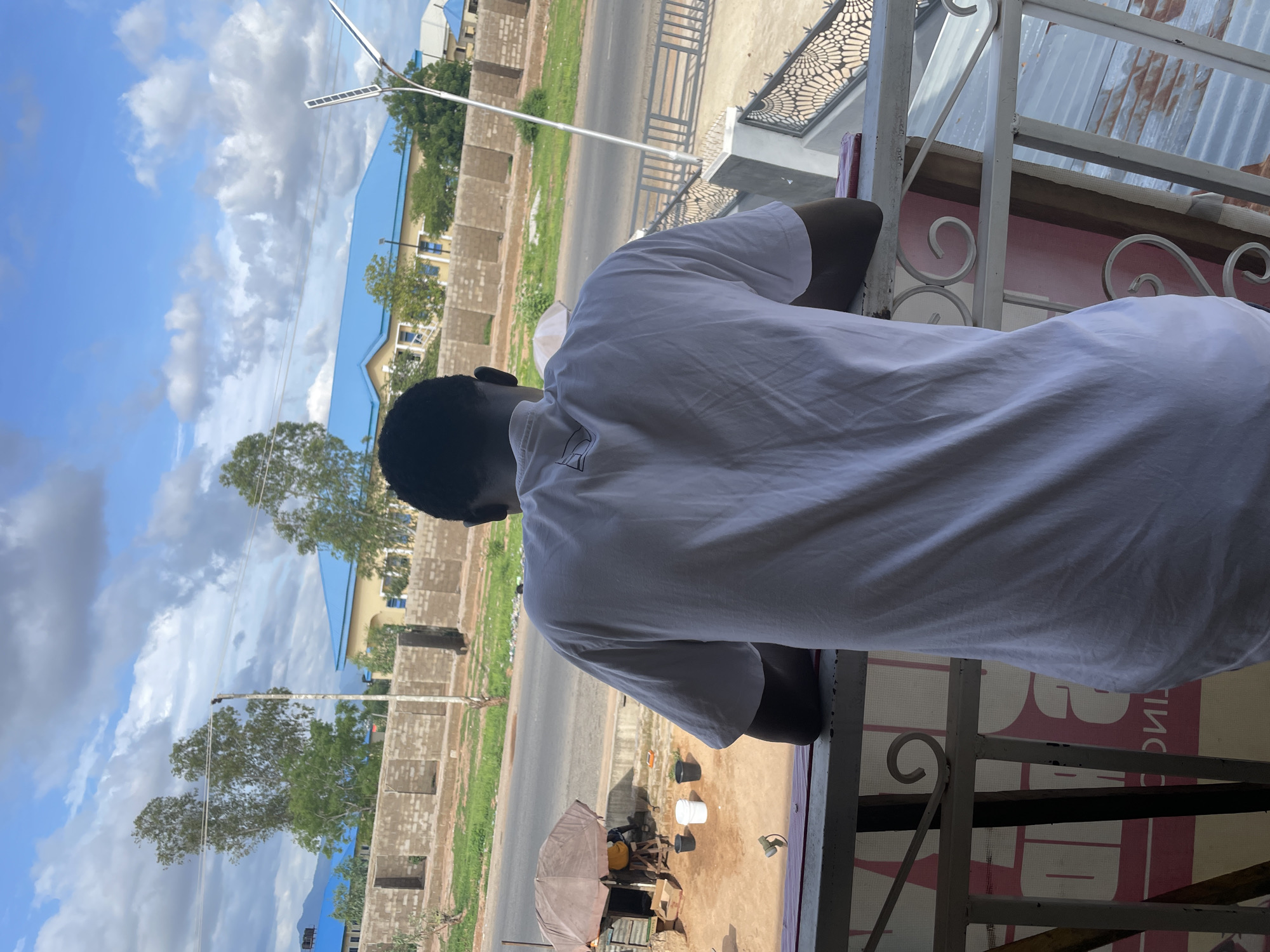 The image depicts an individual observed from behind, leaning on a white metal balcony railing with decorative scrollwork, overlooking a street scene in Jalingo, Nigeria. The person wears a plain white short-sleeved t-shirt featuring a small dark graphic near the back collar. Below the railing, a partial red and white sign is visible.
At street level, an asphalt road runs horizontally, bordered by a dirt and grassy verge. Sections of the street are lined with unfinished concrete block walls. On the left, a vendor's setup is visible under a brown umbrella, where a partially obscured person is positioned behind a large yellow container, with three buckets (two black, one white) on the ground. On the right, a modern street light equipped with solar panels stands behind a grey metal fence featuring a patterned design. In the background, multiple buildings with prominent blue roofs are interspersed among trees and vegetation. The sky is bright blue with scattered white cumulus clouds, and distant hills are faintly discernible on the horizon.