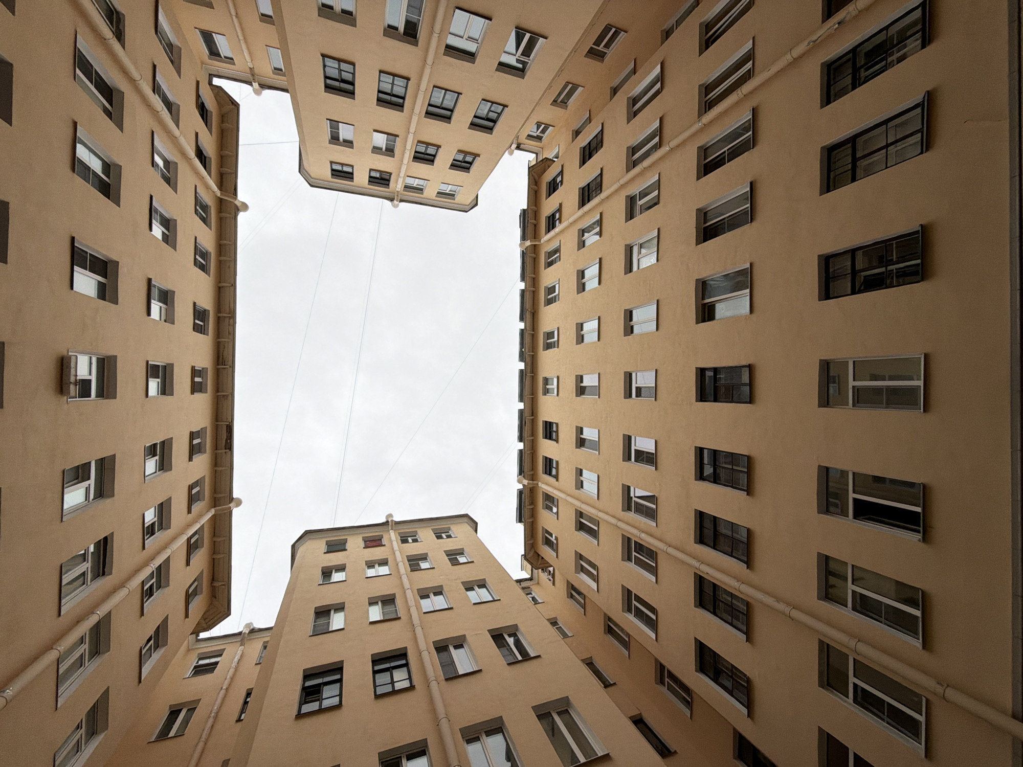A low-angle shot looking up shows buildings surrounding a courtyard in Saint Petersburg, Russia. The buildings are a tan color and feature rows of evenly spaced windows. The sky above the courtyard is overcast. Two thin cables stretch across the open space between the buildings. A downspout is visible on the side of each building.