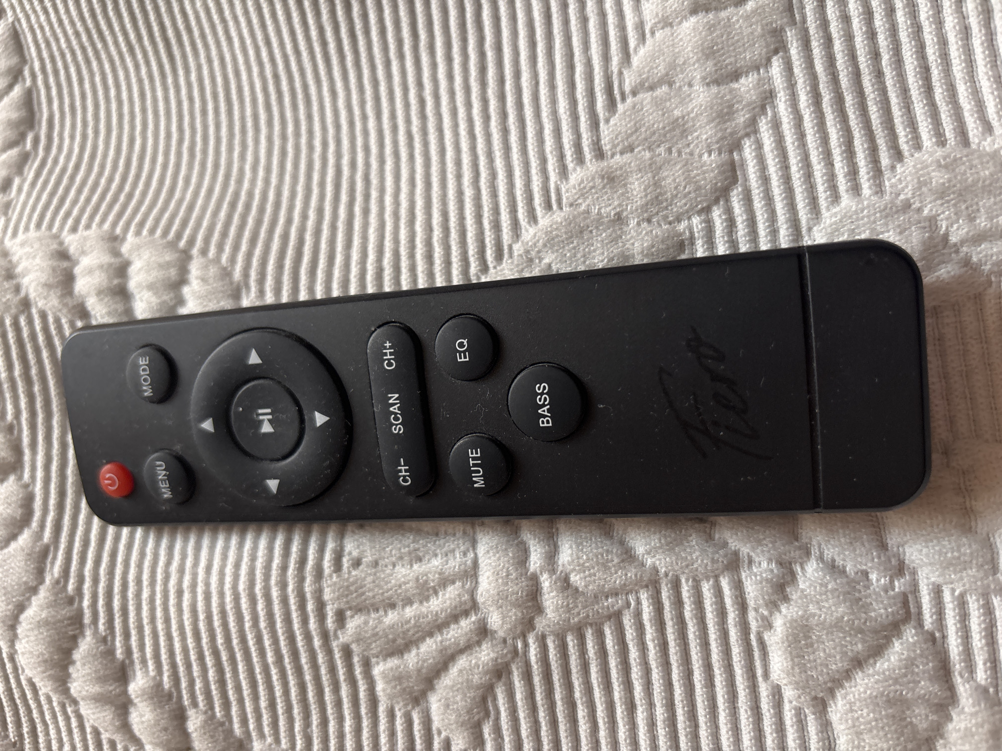 A black remote control with various buttons is lying on a white, textured surface. The remote has a red power button at the top, followed by a menu button and a mode button. Below these is a circular directional pad with play/pause in the center and arrow buttons surrounding it. Further down, there are buttons labeled "CH-", "SCAN", "CH+", "MUTE", "EQ", and "BASS". The brand name "Fiero" is visible near the bottom of the remote. The white surface appears to be a textile, possibly a blanket or tablecloth, with a raised, ribbed pattern. The image was taken in Chernogorsk, Russia.