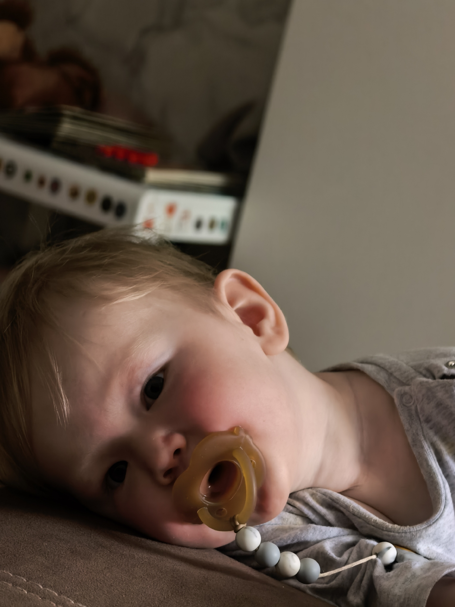 The image depicts a close-up of a baby's face, with the baby lying on its side on a brown surface, possibly a couch or bed. The baby has fair hair and is wearing a gray shirt. A pacifier is in the baby's mouth, and a beaded clip is attached to the shirt. In the background, out of focus, are books and what appears to be a shelf or wall. There is no information or visual cue in the image that indicates its location in the city of Sterlitamak, Russia.