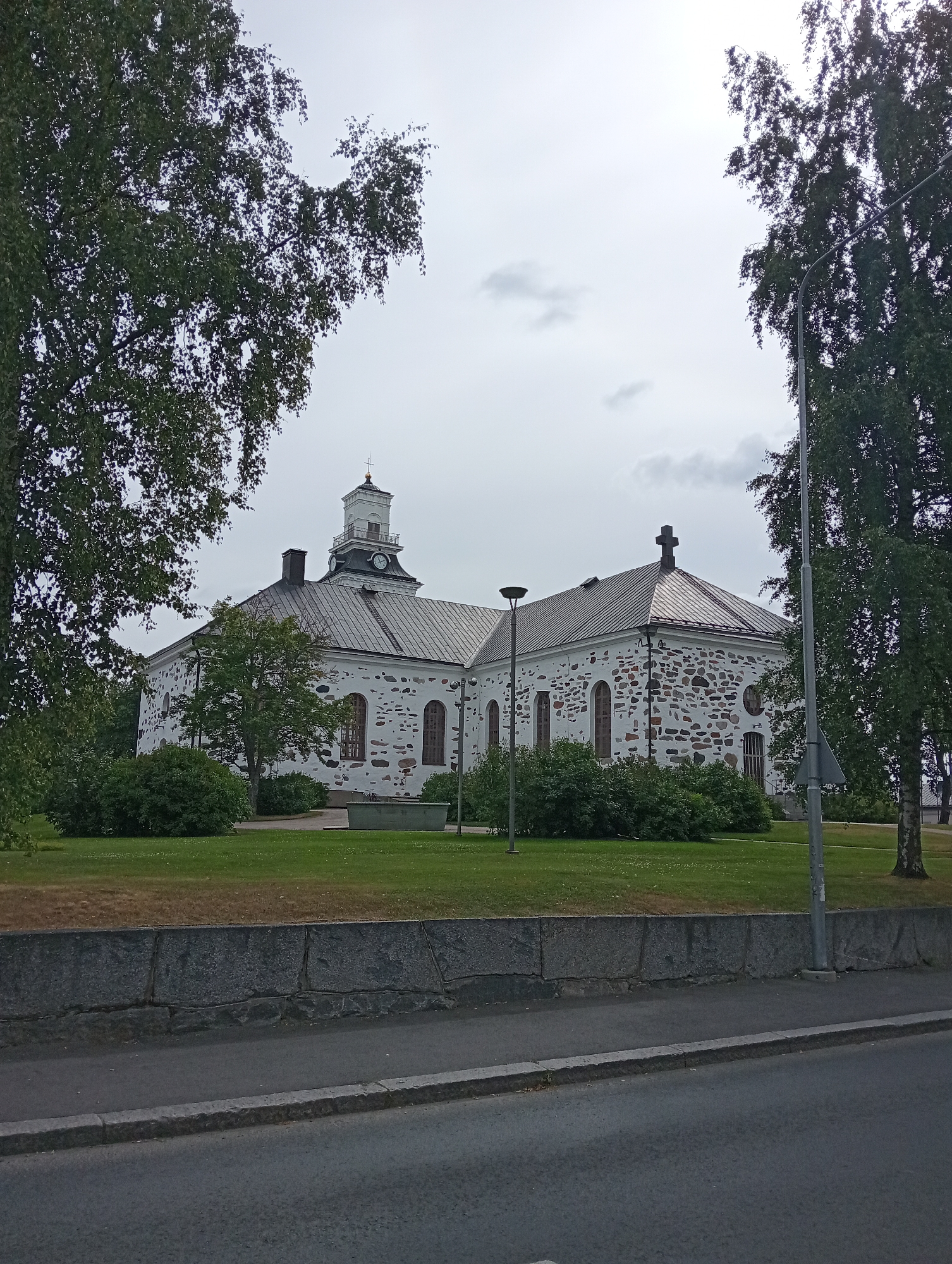 This is a ground-level, eye-level, medium shot of Kuopio Old Church, located in Kuopio, Finland, on a cloudy day. The church is constructed from white stone with darker, irregularly shaped stones embedded in its facade. It features a gray, pitched roof and a prominent clock tower with a white spire and a cross. The church is surrounded by lush green grass, manicured bushes, and several trees, with their leaves providing shade. In the foreground, a gray stone wall separates the church grounds from a paved street. A lamppost and a street sign are visible to the right of the church. There are no people or animals in the frame. The overall scene is calm and serene, suggesting a quiet urban environment.