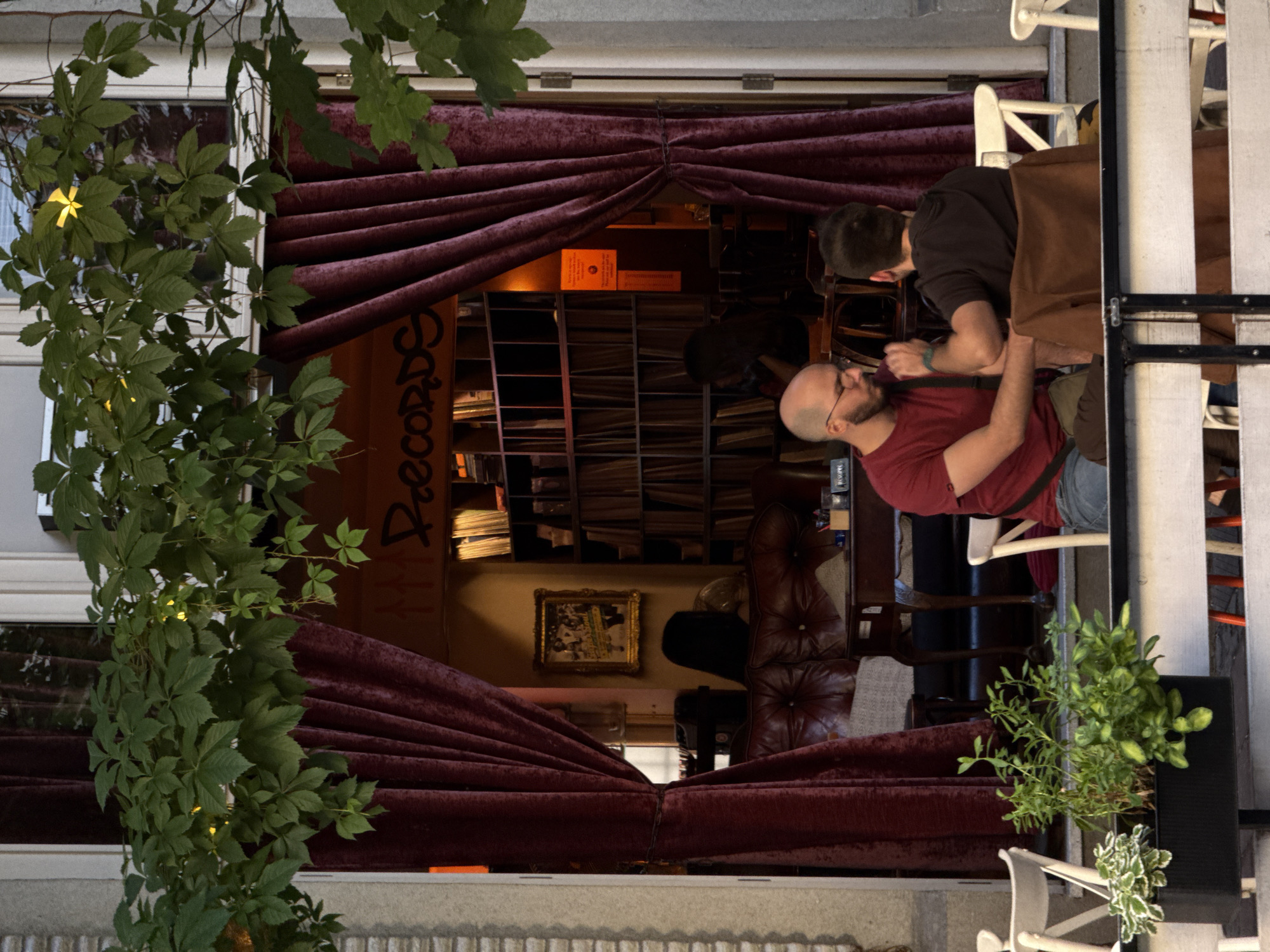 The image depicts the exterior of a record shop in Belgrade, Serbia, as seen through an open doorway draped with burgundy velvet curtains. In the foreground, two men are seated at a table, engaged in conversation. The man on the left, wearing a red t-shirt and glasses, gestures with his hand. The man on the right, wearing a dark t-shirt, faces away from the camera. Beyond them, the interior of the shop is visible, with shelves filled with records and a vintage leather couch. A framed picture hangs on the wall. Green vines with yellow flowers cascade from above the doorway, partially obscuring the view. The overall scene suggests a relaxed atmosphere in an urban setting.