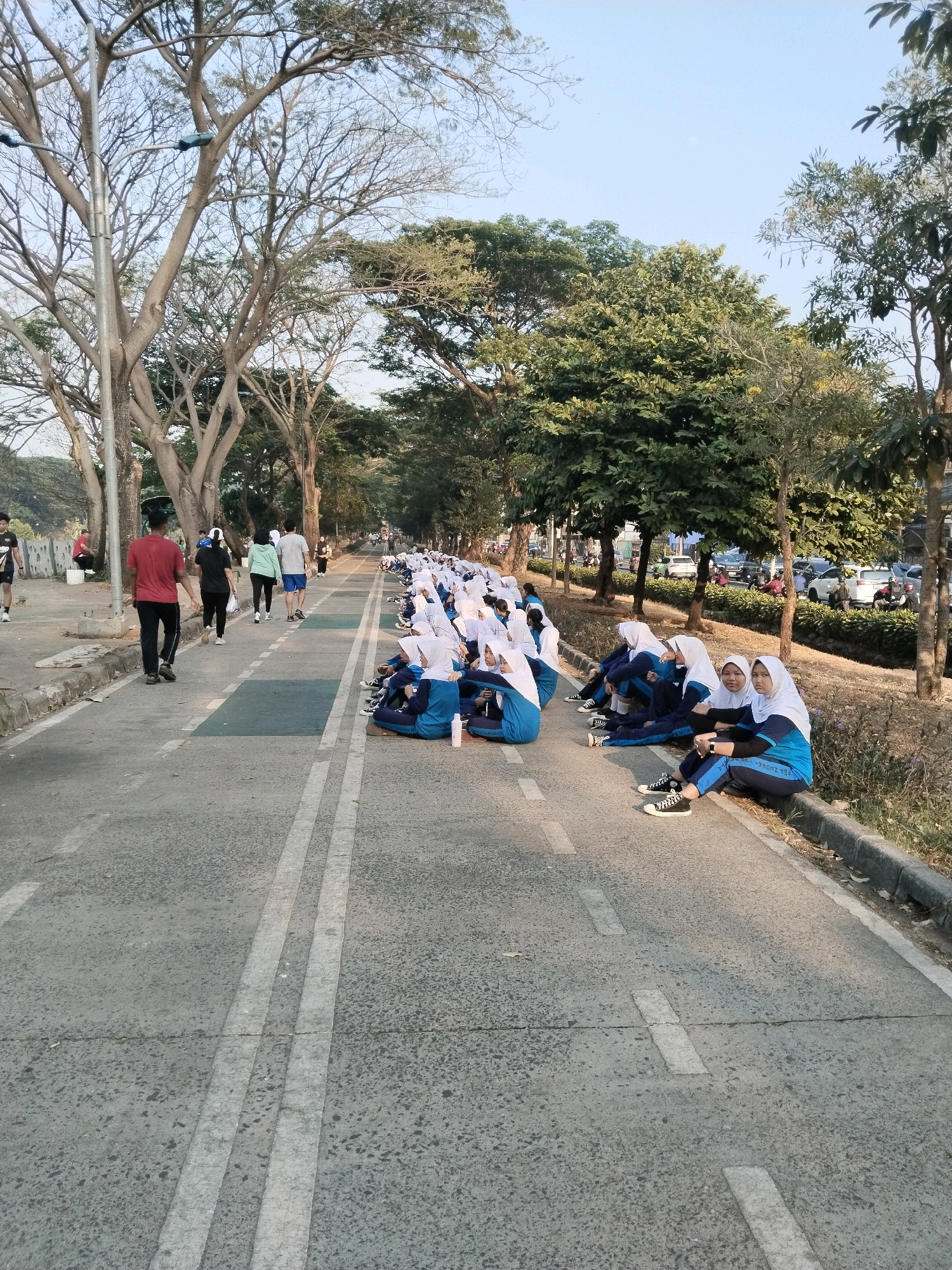 In East Jakarta, Indonesia, an outdoor scene unfolds on a paved road with white dividing lines. A group of young women, dressed in blue uniforms and white hijabs, are sitting on the edge of the road, facing towards the camera. In the background, several individuals are walking and jogging on the road. Trees line the sides of the road, with a clear sky overhead. The road appears to be a path or lane for recreational activities.