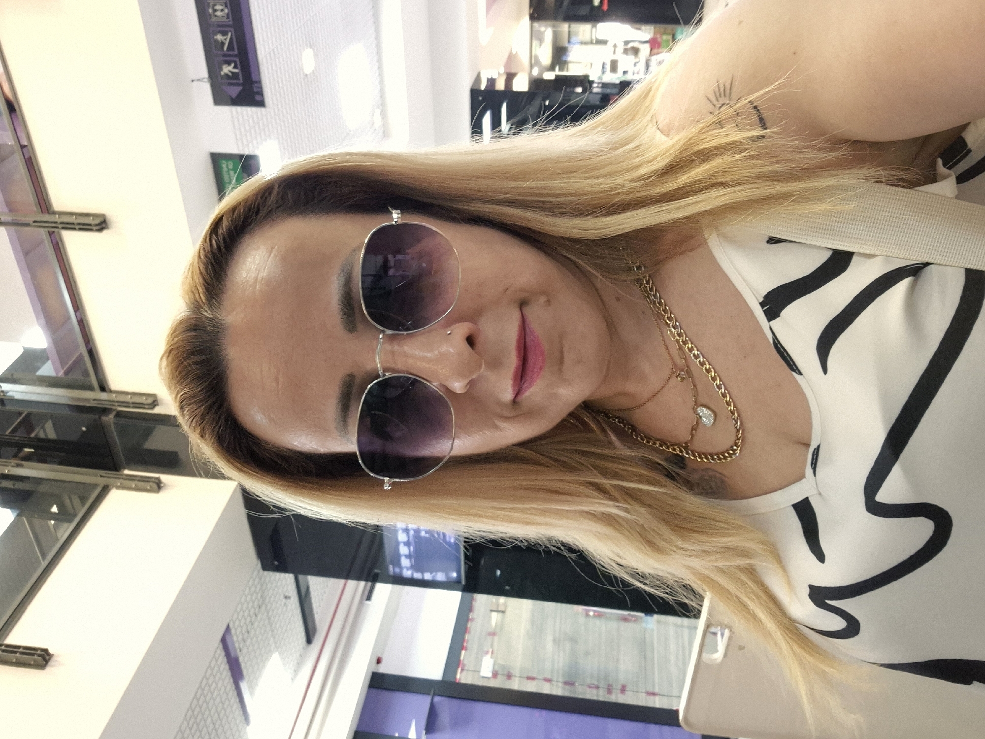 A woman with blonde hair and sunglasses poses for a selfie indoors, possibly in a mall. She is wearing a white top with black abstract stripes and multiple gold necklaces. A portion of a beige strap is visible on her left shoulder, suggesting she is carrying a bag. A small sun tattoo is visible on her right shoulder. The background shows fluorescent lighting and architectural elements of a building.