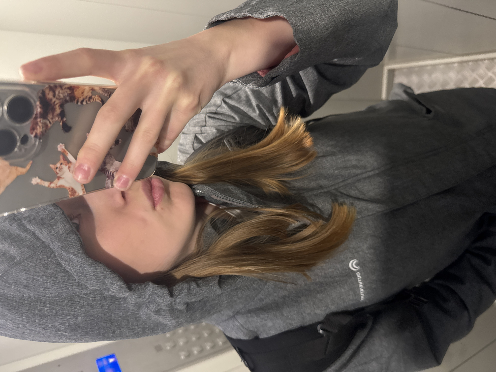 A fair-skinned young person with honey-blonde hair takes a selfie indoors using a smartphone with a cat-themed case. The person wears a gray hooded sweatshirt and a black jacket. The location appears to be a building with white walls. The presence of what seems to be an elevator panel with a blue-lit button suggests the location is in an elevator. The city of Chaikovsky, Russia, cannot be determined.
