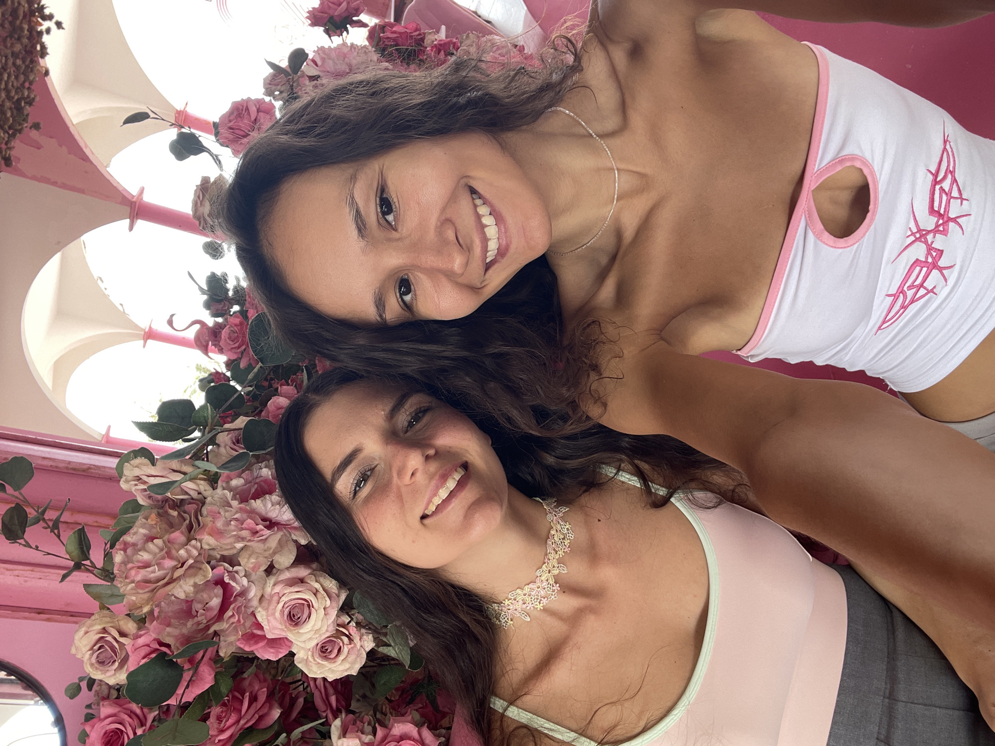 Two young women are taking a selfie in front of a large floral arrangement of pink and white roses. The woman on the left has long, dark brown hair and is wearing a light pink tank top with a mint green trim and a delicate floral choker necklace. The woman on the right has long, curly dark brown hair and is wearing a white strapless top with pink trim and a cutout detail on the chest. She is also wearing a simple beaded necklace. The background includes pink architectural elements and white arches, suggesting an outdoor or semi-outdoor setting, possibly a cafe or event space. The overall atmosphere is bright and cheerful, with a focus on the women and the abundant flowers. The image does not provide any direct information about the specific location in Tien Thanh Ward, Vietnam.