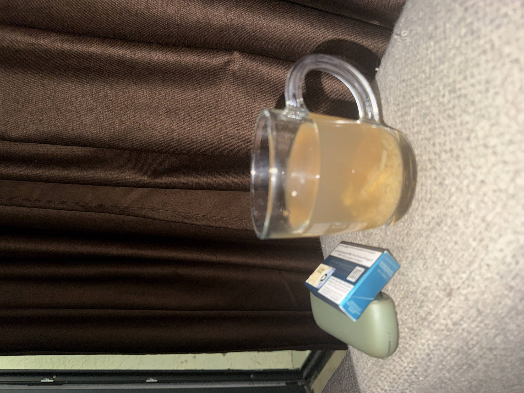 An indoor, close-up, eye-level, medium shot shows a clear glass mug filled with a light-colored liquid and some solid particles, possibly food or tea. The mug has a handle and is placed on a textured, light-colored surface, possibly a cushion or blanket. To the left of the mug, a small, blue and white box, likely a pack of cigarettes, is propped against a light green, oval-shaped object. In the background, dark brown, pleated curtains obscure the view. The overall lighting suggests a dimly lit environment. The scene is located in Tbilisi, Georgia.