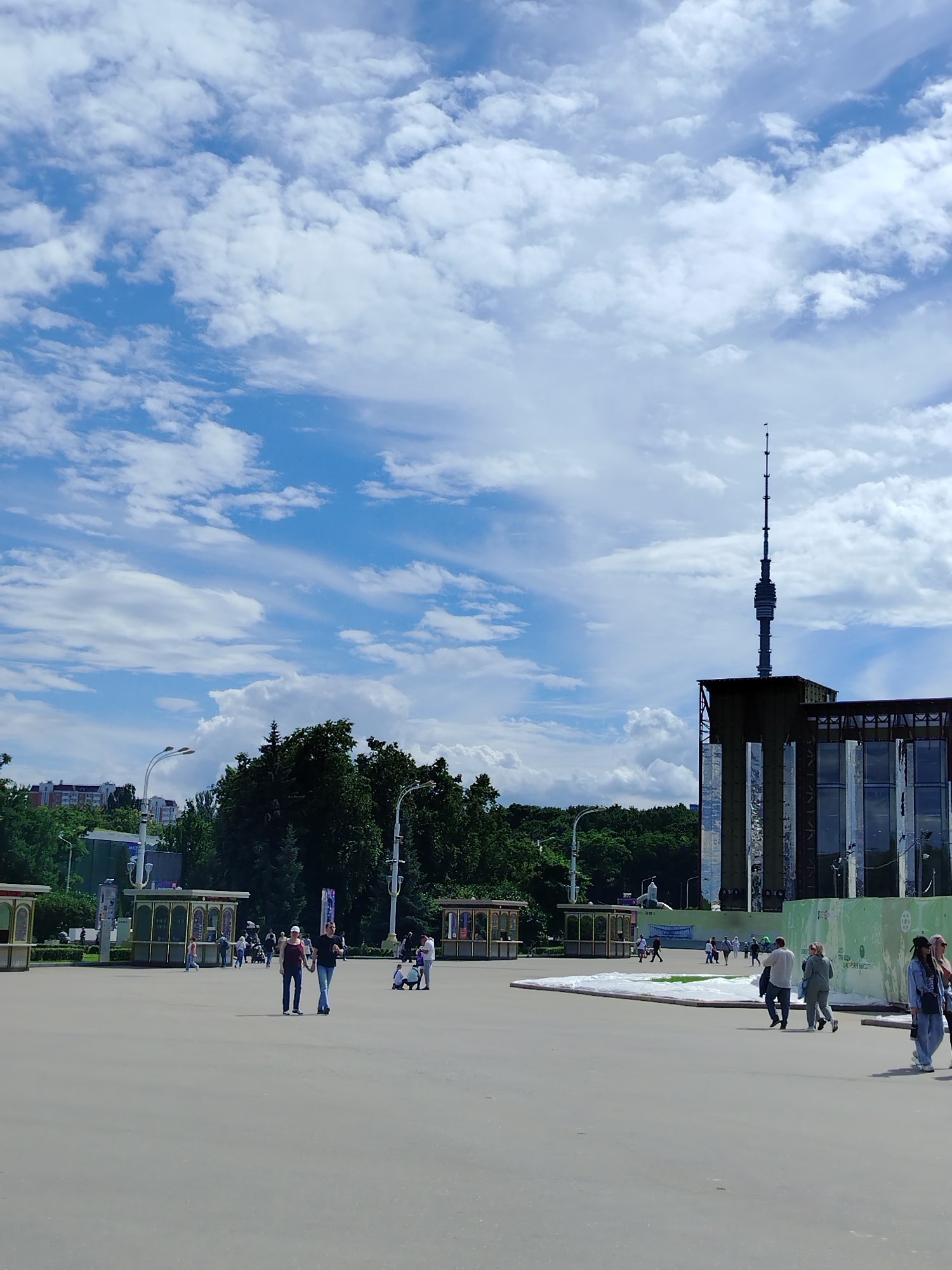 The image depicts a wide, paved outdoor plaza with several people walking in various directions. In the background, a tall television tower, the Ostankino Tower, dominates the skyline, with a modern building featuring large glass windows partially obscuring its base. To the left of the tower, a line of green trees forms a natural boundary, and beyond that, apartment buildings are visible. The sky is a vibrant blue, populated by scattered white clouds. Several small, enclosed kiosks with transparent roofs are dotted around the plaza. The overall scene suggests a public space in Moscow, Russia, on a bright, cloudy day, likely a park or exhibition ground.
