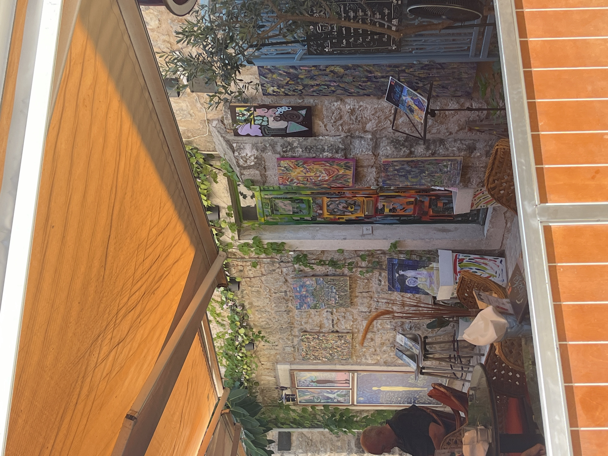 The image shows an outdoor art gallery or display in Budva, Montenegro. The art is hung on a stone wall covered in vines. An orange awning provides shade. A cafe or seating area is visible below, with a person sitting at a table.