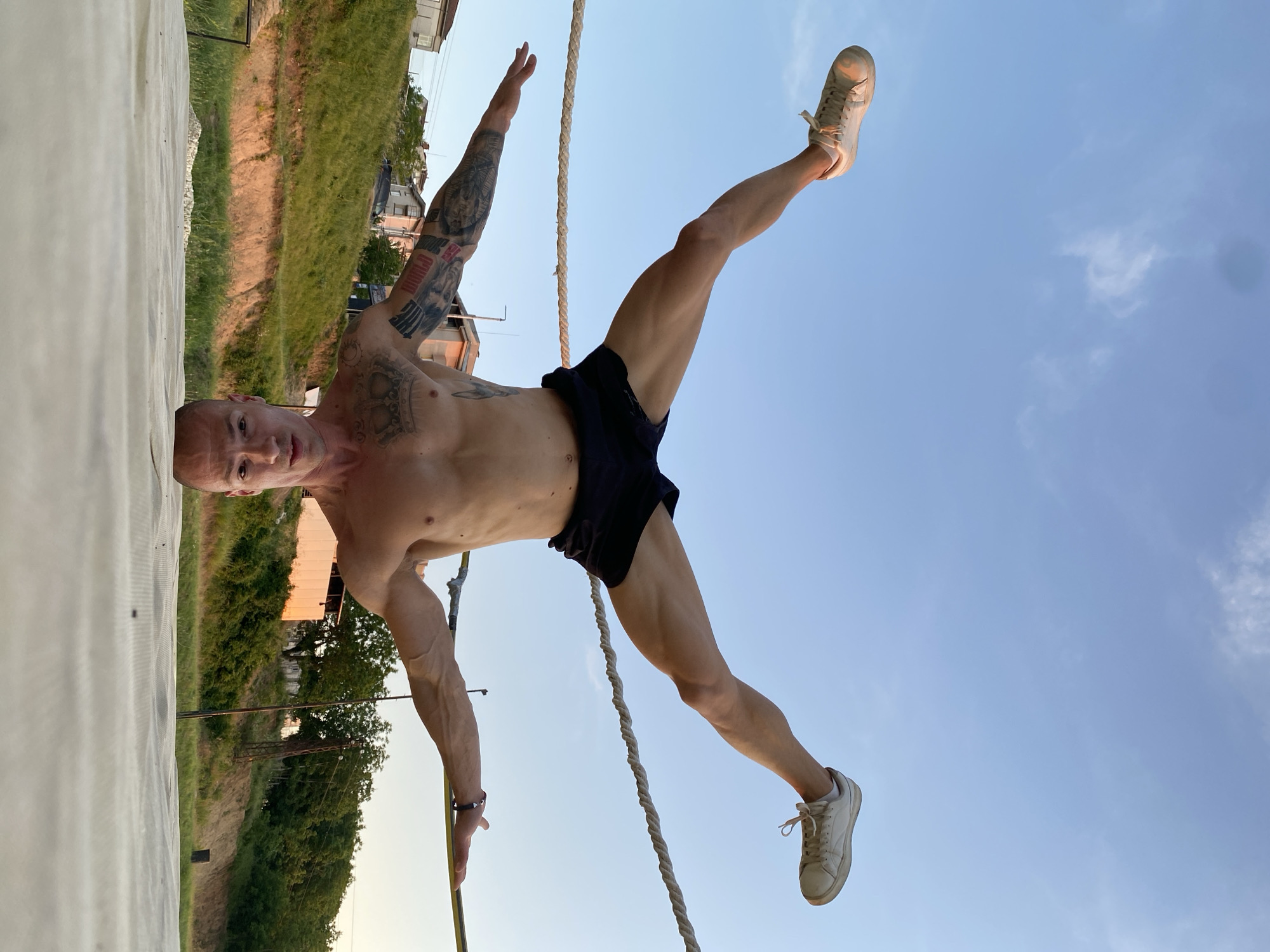 An adult male is performing a handstand with his head on a white mat. His body is upside down, with his legs spread apart and raised above him, each wearing a white sneaker. His arms are extended outwards, with his hands on a thin yellow line. He has tattoos on his arms and torso, and is wearing black shorts. The background shows a grassy hill with houses and trees. The sky is blue with some clouds. The image appears to be taken at dusk. The location is not specified as Pleven, Bulgaria.