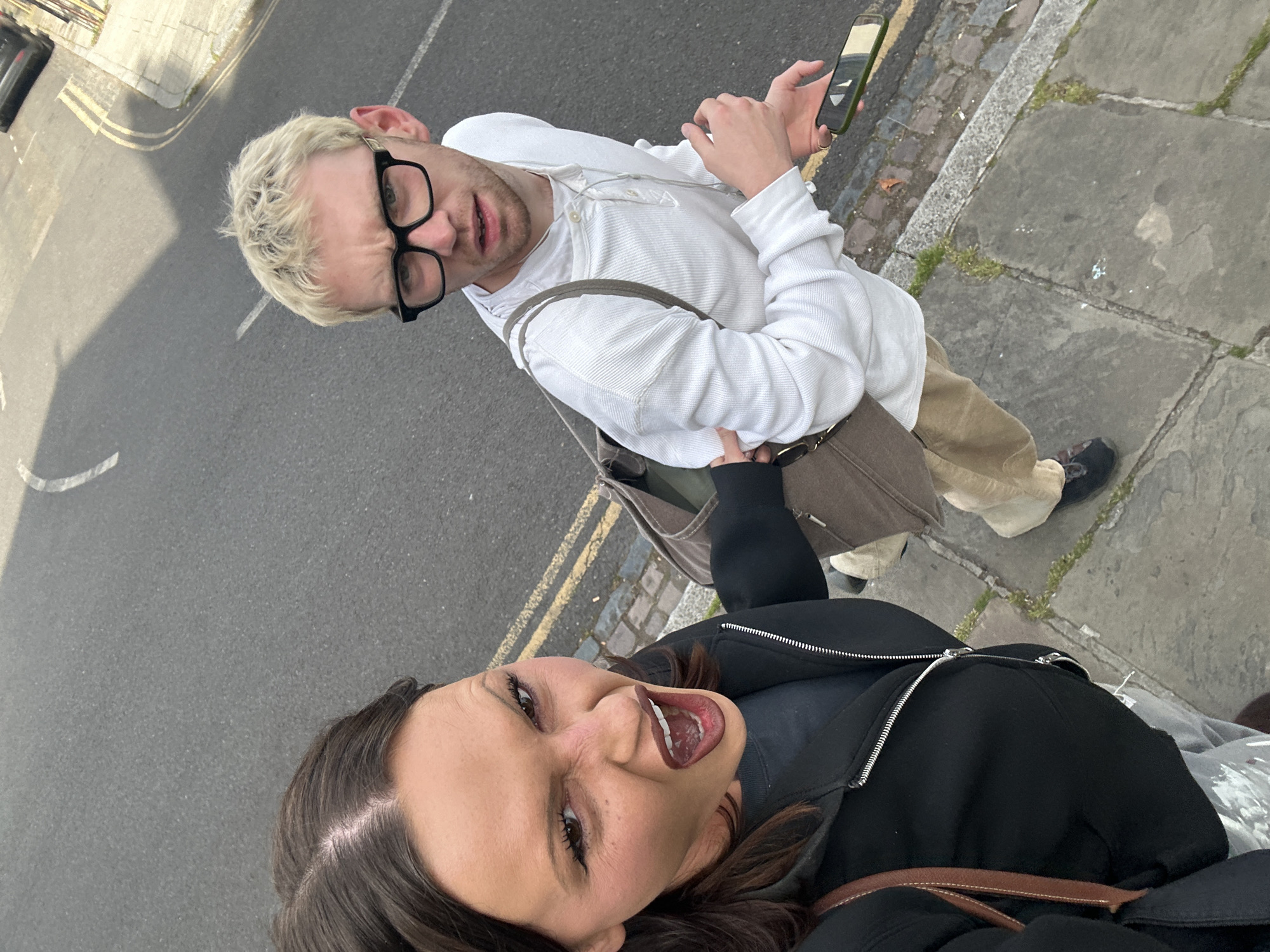 Two individuals are captured in a selfie taken outdoors. The person in the foreground, presumably taking the photo, is a woman with dark brown hair, wearing a black jacket and a brown shoulder bag. Her mouth is open in a wide expression. Behind her, a fair-skinned man with bleached blonde hair and glasses stands wearing a white top, beige trousers, and a brown bag. He is holding a mobile phone in his hands. They are standing on a gray pavement next to a gray street with yellow painted lines, likely in an urban setting. The scene suggests a location in London, United Kingdom, given the typical British street aesthetics.