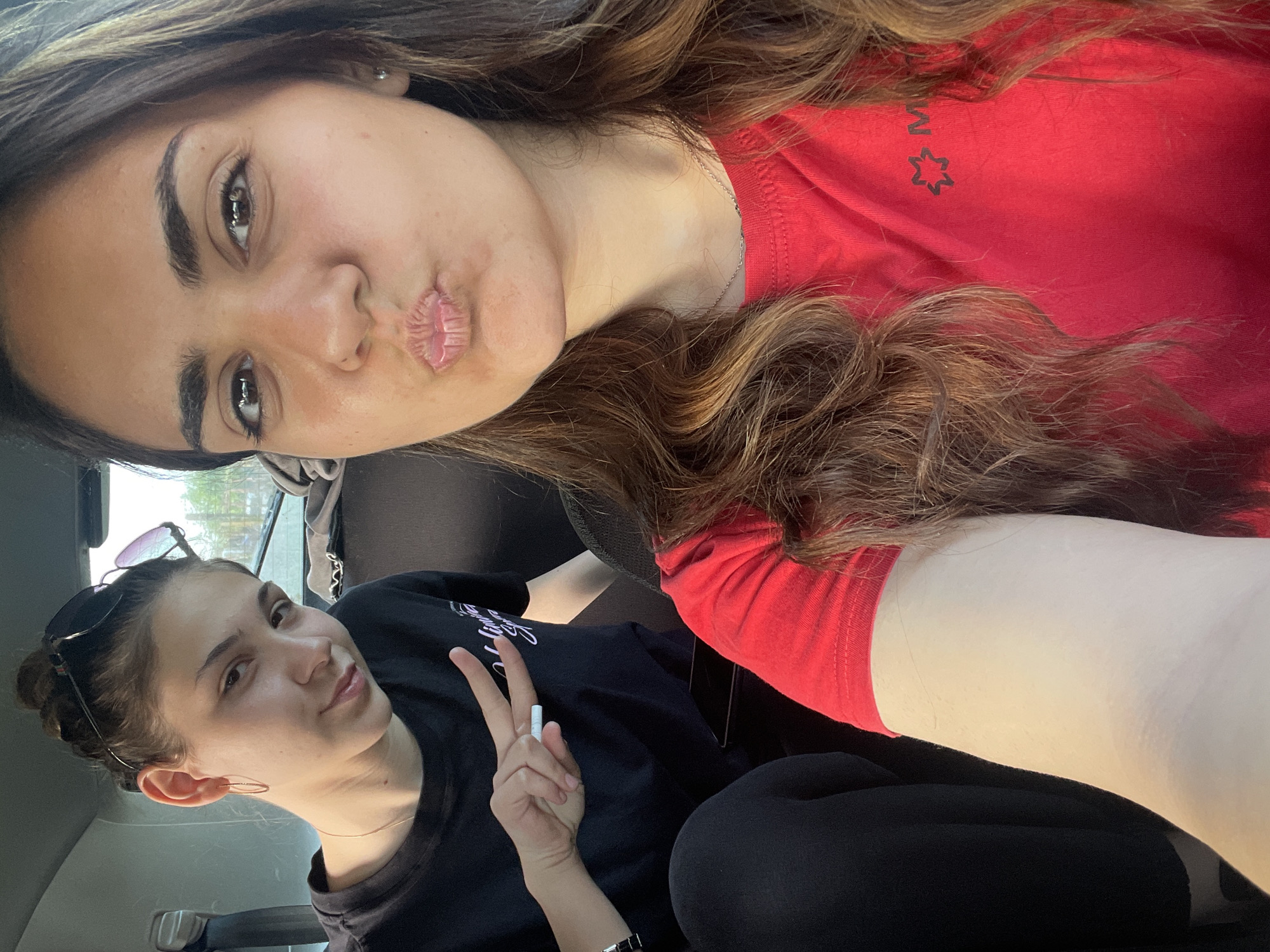 Two women are in the backseat of a car. The woman in the foreground is wearing a red t-shirt with a logo on the chest. The woman in the background is wearing a black t-shirt with text and a logo. She is wearing sunglasses on her head and making a peace sign with her left hand.