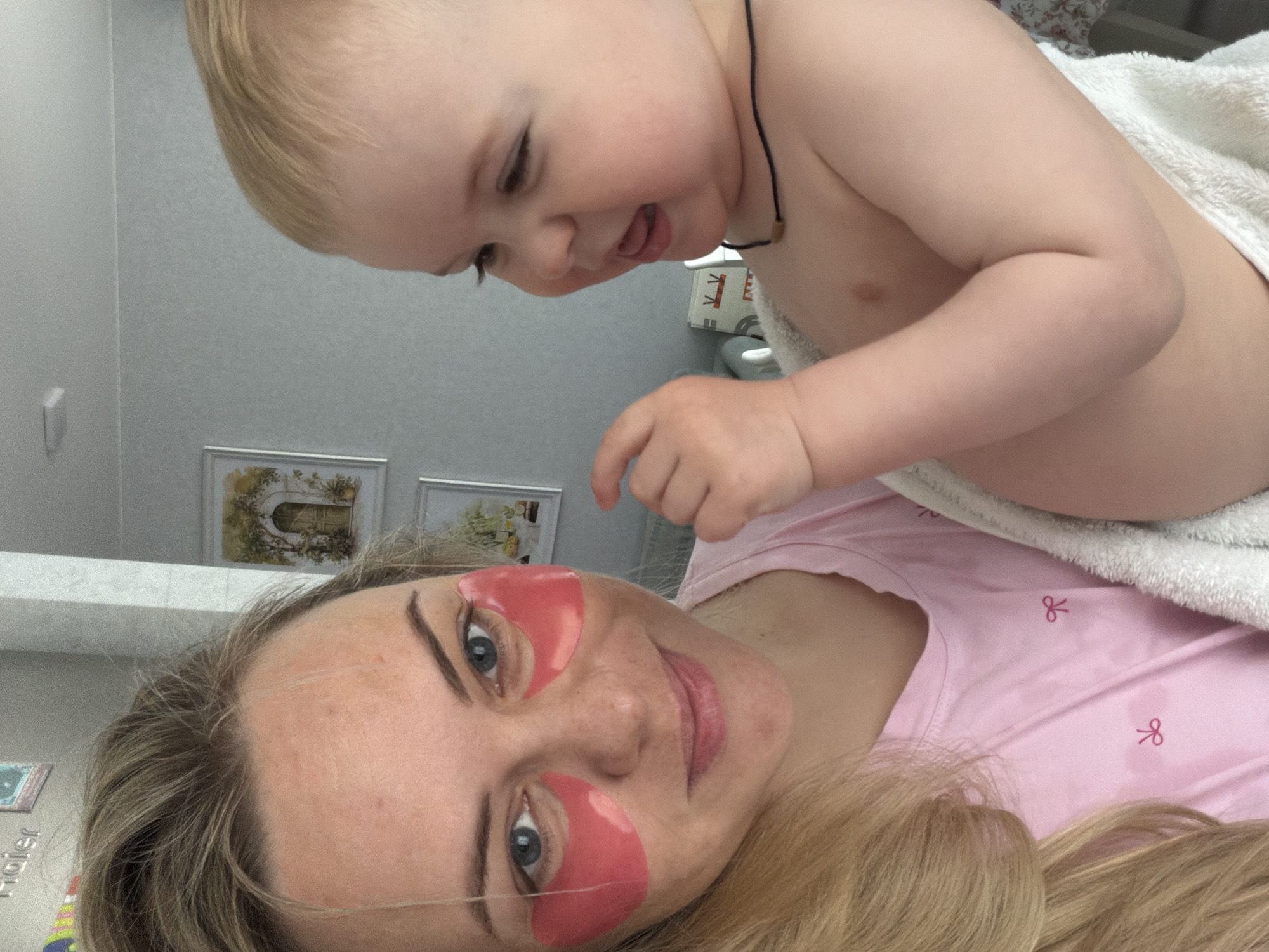 A selfie shot in an indoor setting shows a woman and a young child. The woman is lying down, wearing pink eye patches, and has blonde hair. She is wearing a pink top with small bow designs. The child is fair-skinned with blonde hair and is partially wrapped in a white towel. The child has a necklace with a dark cord. The background includes framed pictures on a wall, a bedside table with items on it, and neutral-toned walls. It is not possible to determine if the location is Votkinsk, Russia from this image.