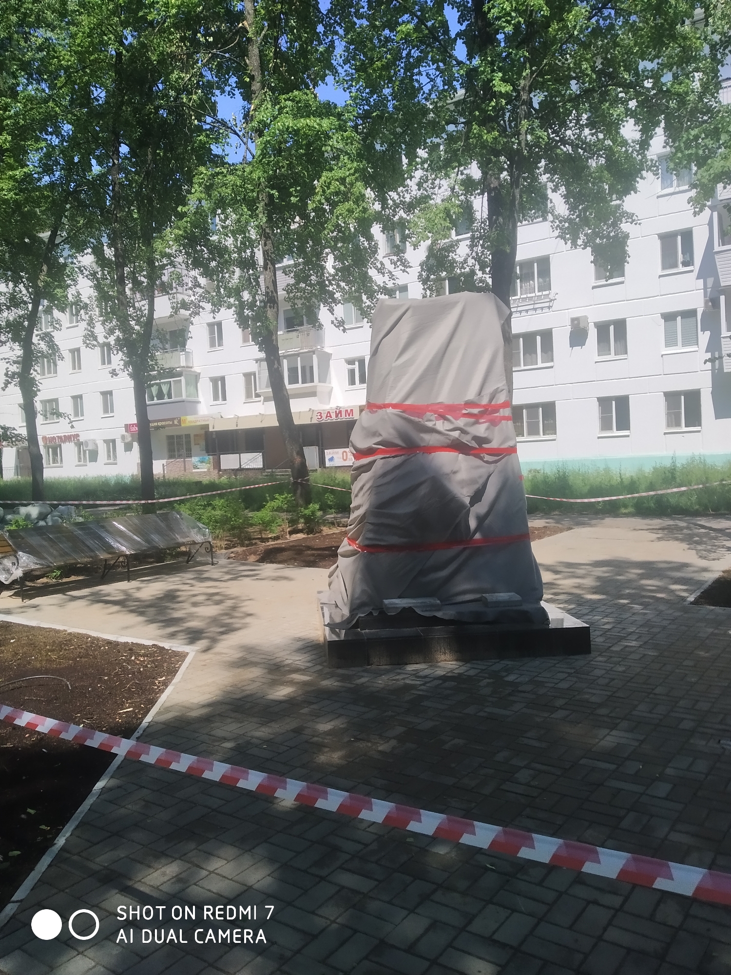 The image captures an outdoor urban scene, likely a public square or park, in Chaikovsky, Russia, on a bright, sunny day. In the foreground and midground, a tall, columnar monument or sculpture stands centrally, completely covered by a light grey tarp. Red tape is wrapped around the tarp in two horizontal bands to secure it. The monument rests on a multi-tiered, dark-colored base.
To the left of the covered monument, a park bench is visible, partially draped with clear plastic sheeting, suggesting recent installation or protection. Patches of bare earth or mulch surround the paved pathways made of grey rectangular pavers. A red and white striped caution tape runs across the lower foreground, indicating a restricted area or ongoing work. Several tall trees with full green foliage are scattered throughout the scene, casting dappled shadows on the ground and creating a verdant canopy against a clear blue sky.
In the background, a large, white, multi-story apartment building dominates, featuring numerous windows and some balconies. The ground floor of this building houses several commercial establishments. Visible Cyrillic text on the shop signs includes "НОТАРИУС" (Notary) on the far left, fragments like "FAB...", "...АЙМ КРАСОТЫ" (Beauty Salon/Realm of Beauty), and "КВАДРАТ..." (Square/Quadrant). Most prominently, a sign reads "...ЗАЙМ" (Loan/Pawnshop) with "ЗАЙМ 0%" (0% Loan) underneath. An air conditioning unit is mounted on the building's exterior. No people are visible in the scene, which appears to be in a temporary state of readiness or renovation, possibly awaiting the unveiling of the covered monument. The image bears a "SHOT ON REDMI 7 AI DUAL CAMERA" watermark at the bottom left.
