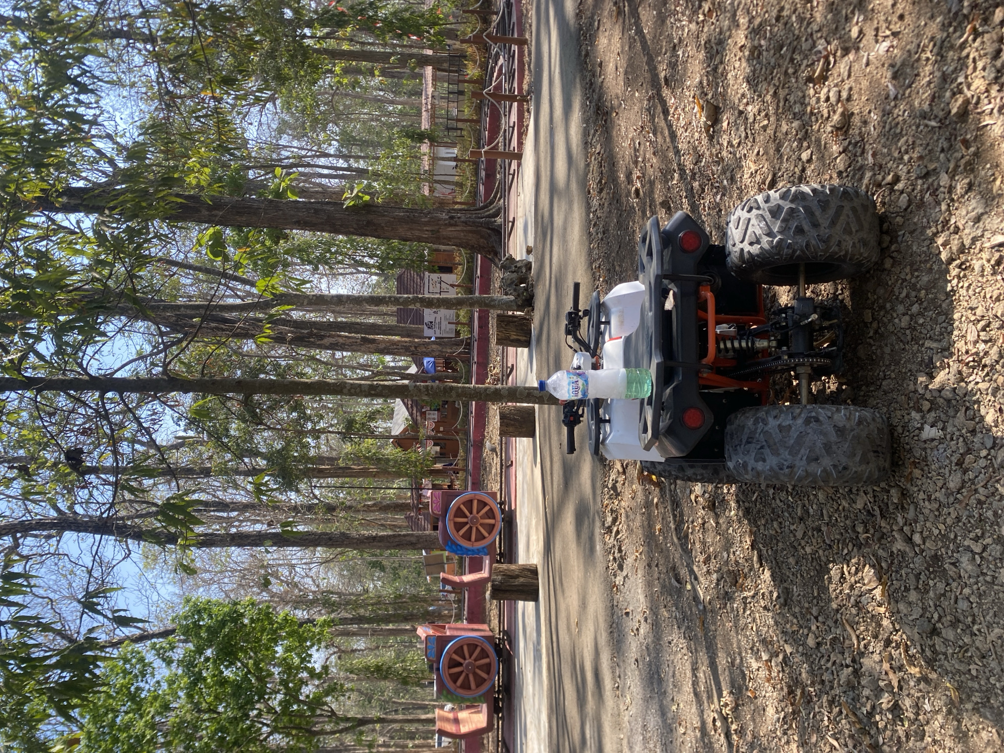 In the city of Ketileng, Indonesia, an all-terrain vehicle (ATV) is parked on a gravelly path in a wooded area. The ATV is white with an orange frame and black tires. A bottle of water sits on the back of the ATV. In the background, there are several trees and a small amusement park with train-like rides. The sky is clear and blue.