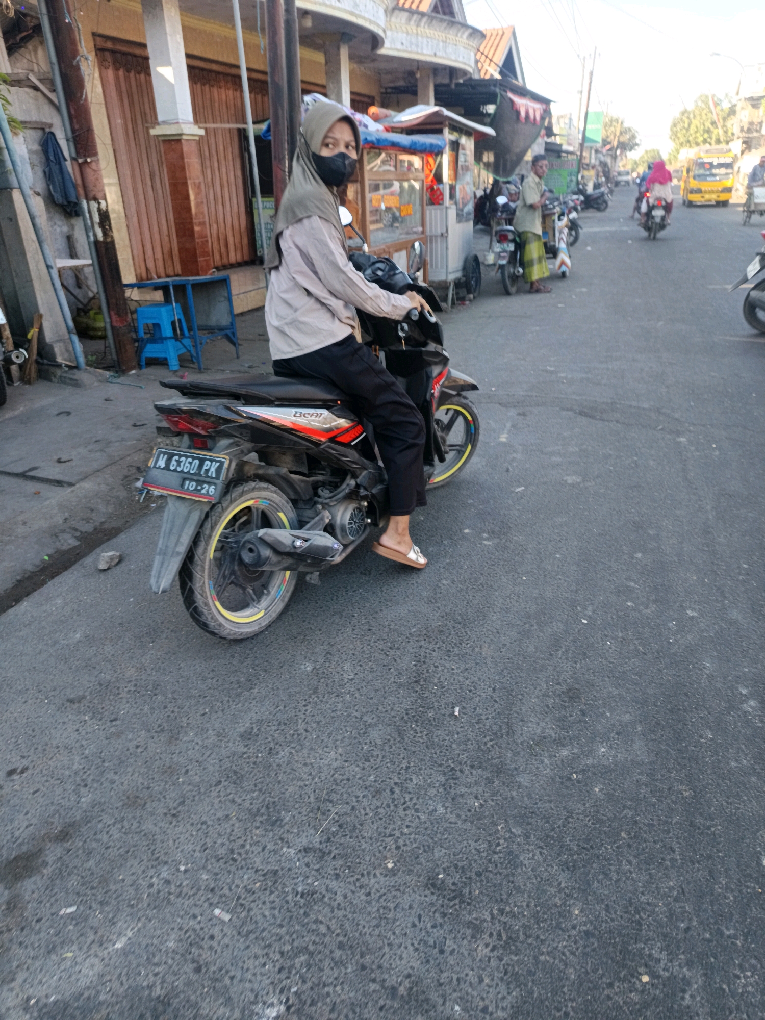 A person wearing a hijab and a face mask is sitting on a motorcycle in the city of Blega, Indonesia. The person is wearing a light-colored long-sleeved shirt and dark pants, with brown sandals. The motorcycle is black and red, with a license plate that reads "M 6360 PK 10-26". The background shows a street scene with buildings, other motorcycles, and a yellow truck. The image is taken during the day, with bright sunlight casting shadows. The street appears to be paved with asphalt.