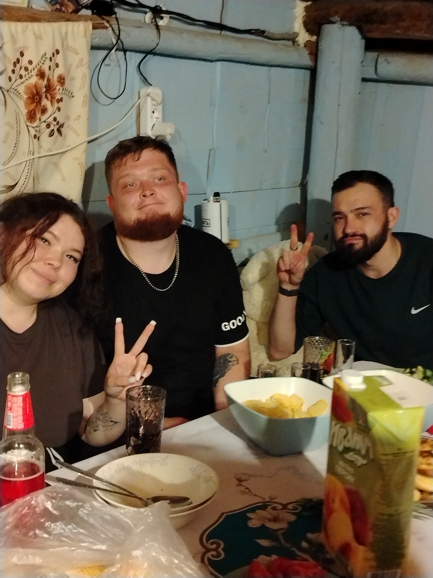 Three individuals are seated at a table laden with food and beverages in what appears to be a home in Гожан, Russia. A young woman with dark curly hair is on the left, making a peace sign. To her right is a man with a reddish beard and a gold chain. He is wearing a black t-shirt. The man on the right also displays a peace sign.
The table holds a bottle of red soda, a glass, plates, silverware, a plastic bag containing food, a bowl of chips, and a carton of juice. The room features paneled walls painted light blue. A flowered curtain hangs on the wall to the left. The image is taken indoors. The table has a floral patterned tablecloth. There are dark wood rafters at the top of the frame.