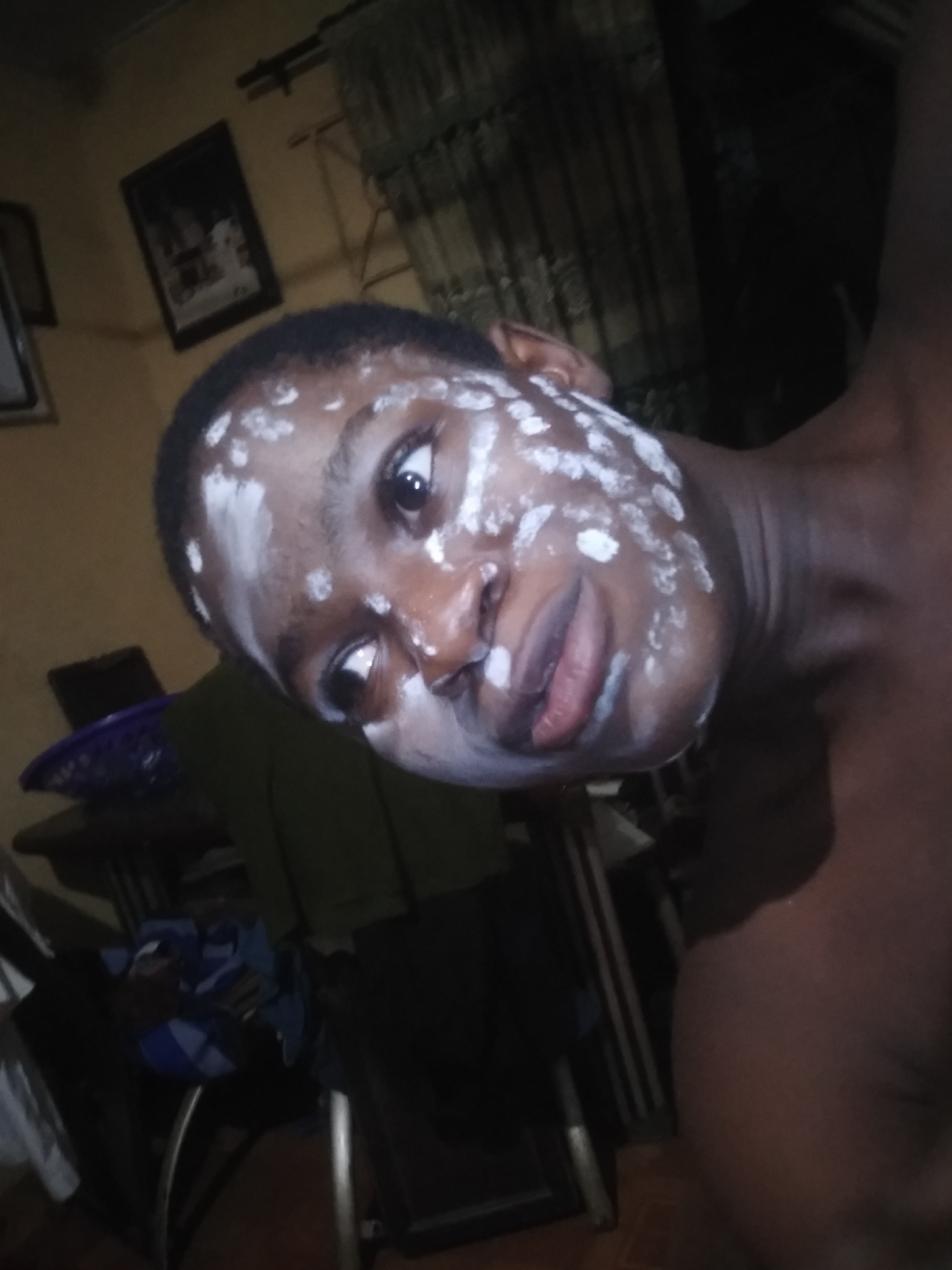 A dark-skinned person with white cream or paste dotted on their face is looking at the camera. The person appears to be shirtless. The background is a dimly lit room with a curtained window, pictures on the wall, and furniture partially visible. There is no specific, identifiable information to suggest the location is Shangisha, Nigeria.