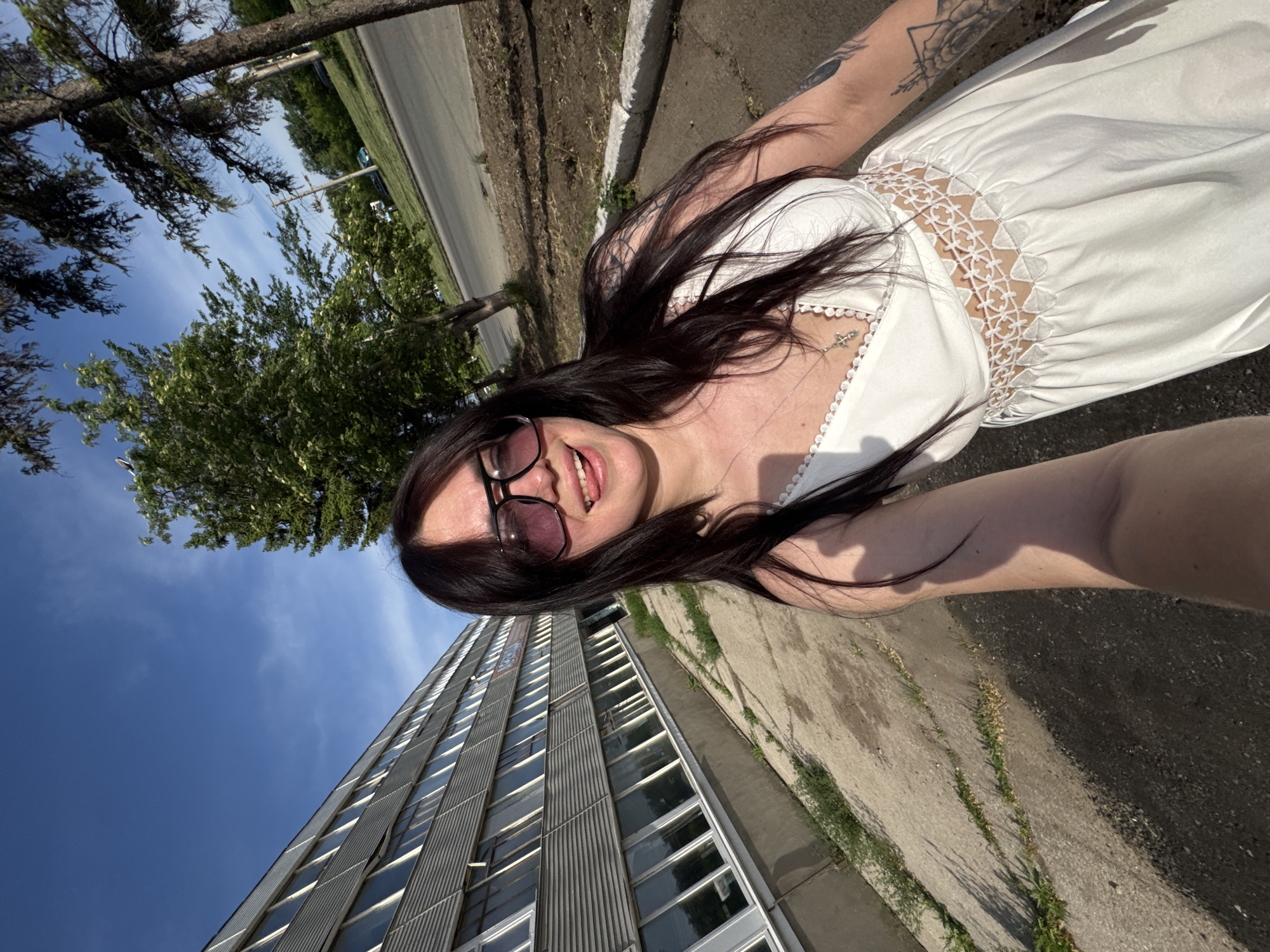 A selfie of a woman wearing a white dress and sunglasses, with a large gray building and blue sky in the background. The woman has dark hair and fair skin, and she is wearing a cross necklace. Her tongue is slightly out of her mouth. The building has a modern design with rows of windows, and it is located on a city street. The ground is concrete with patches of grass growing through cracks. There is also some greenery and trees visible in the background. The location appears to be an urban area.
