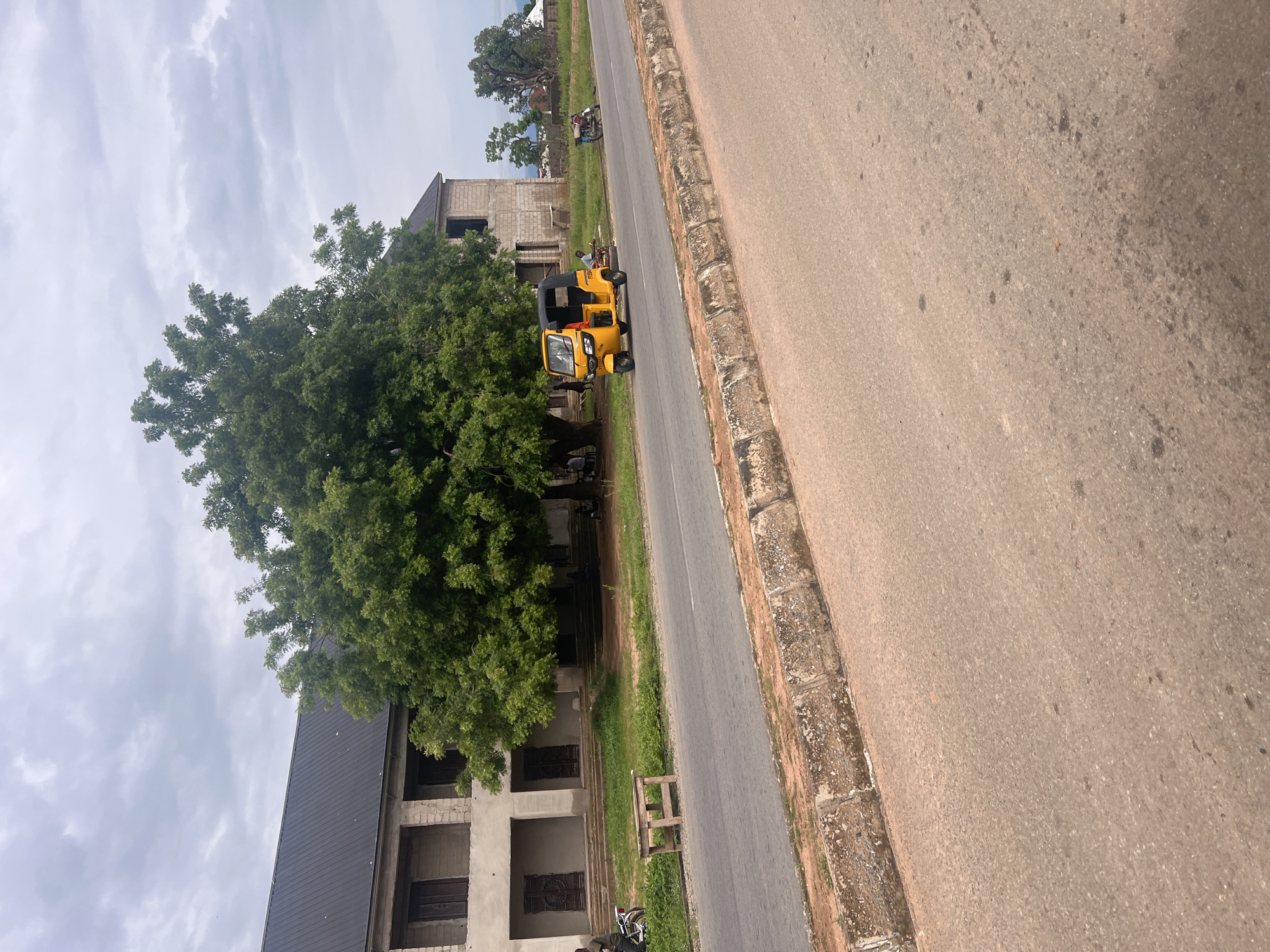 The image shows a scene in Jalingo, Nigeria, with a road running diagonally across the frame. On the left side of the road, there is a large, lush green tree, and a partially constructed building made of concrete blocks. A yellow, three-wheeled vehicle, often called an "okada" or "Keke," is parked on the side of the road next to the tree. A person is visible in the driver's seat of the Keke. Further down the road, another motorcycle is parked. The building appears to have a dark, sloped roof. The road itself is paved with asphalt, and there is a curb on the right side. The sky is overcast with clouds. The overall impression is of a typical day in a town in Nigeria, possibly a residential or commercial area under development. There is no visible text in the image.