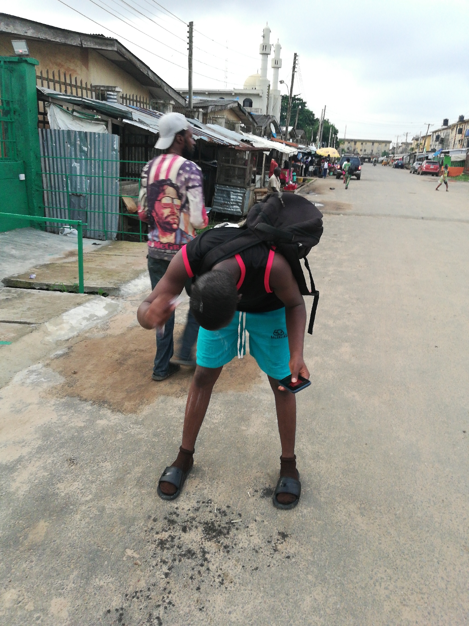The image shows two individuals on a street in an urban environment. The foreground subject, a person with dark skin, is bent over looking at a mobile phone held in their right hand. They are wearing a black top with pink straps, turquoise shorts, socks, and sandals, and have a black backpack on. In the background, another individual with dark skin is walking away. They are wearing a grey cap, a t-shirt with a graphic print, and jeans. Buildings and structures line the street, including a mosque with two minarets in the distance. Other details visible include a car, a bicycle, and additional people farther down the street. The street itself is paved, and there are some puddles of water on the ground. The sky is overcast.