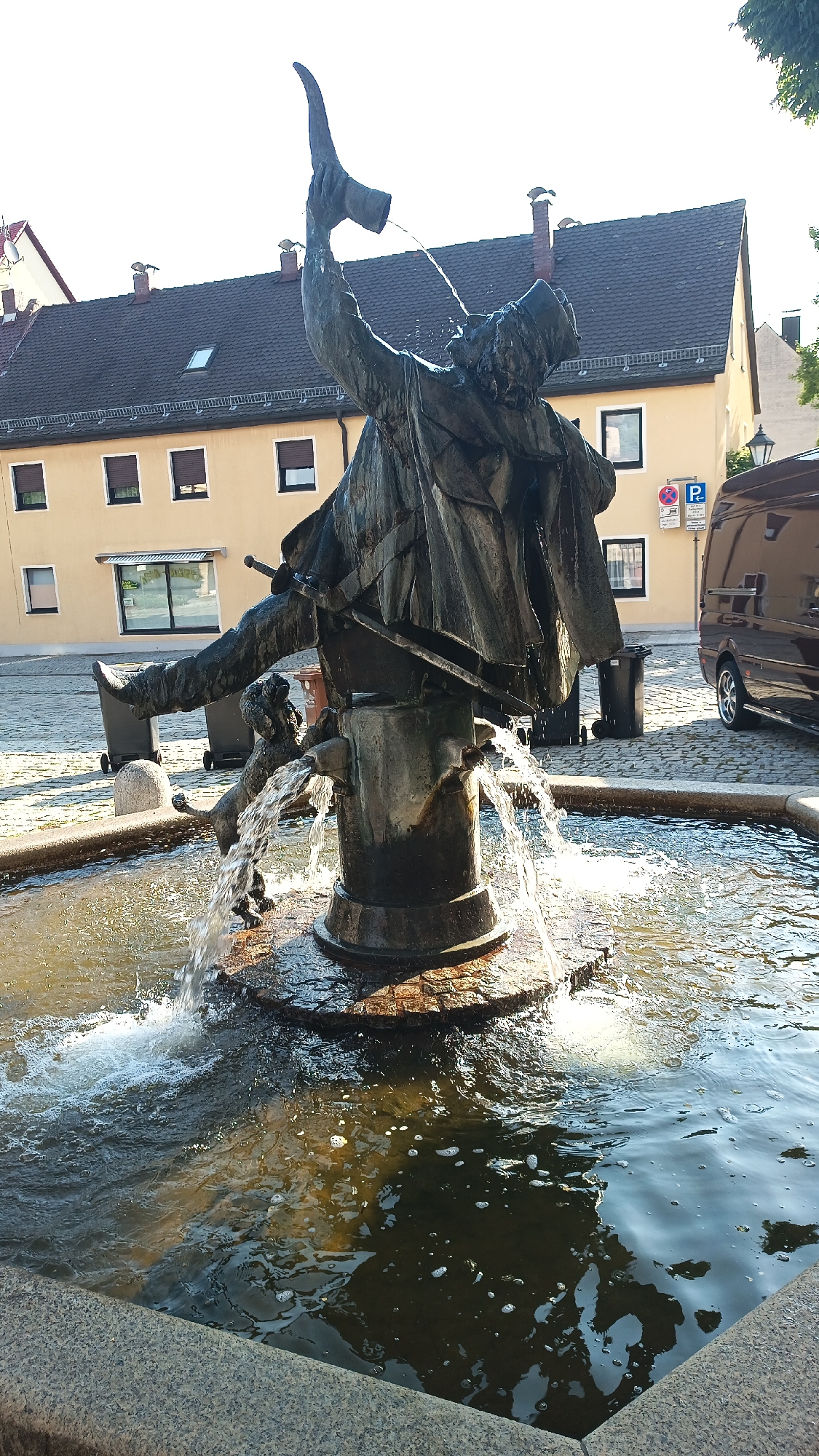 A bronze fountain sculpture depicts a man in a hat and cape, raising a horn from which water pours, in the town square of Unknown, Unknown. A small dog is positioned at the base of the sculpture, also spraying water. The fountain is surrounded by a stone basin filled with water, with a cobblestone pavement beyond. In the background, a yellow building with dark tiled roofs and windows is visible, as are trash bins and a dark van. The sky is clear and bright.