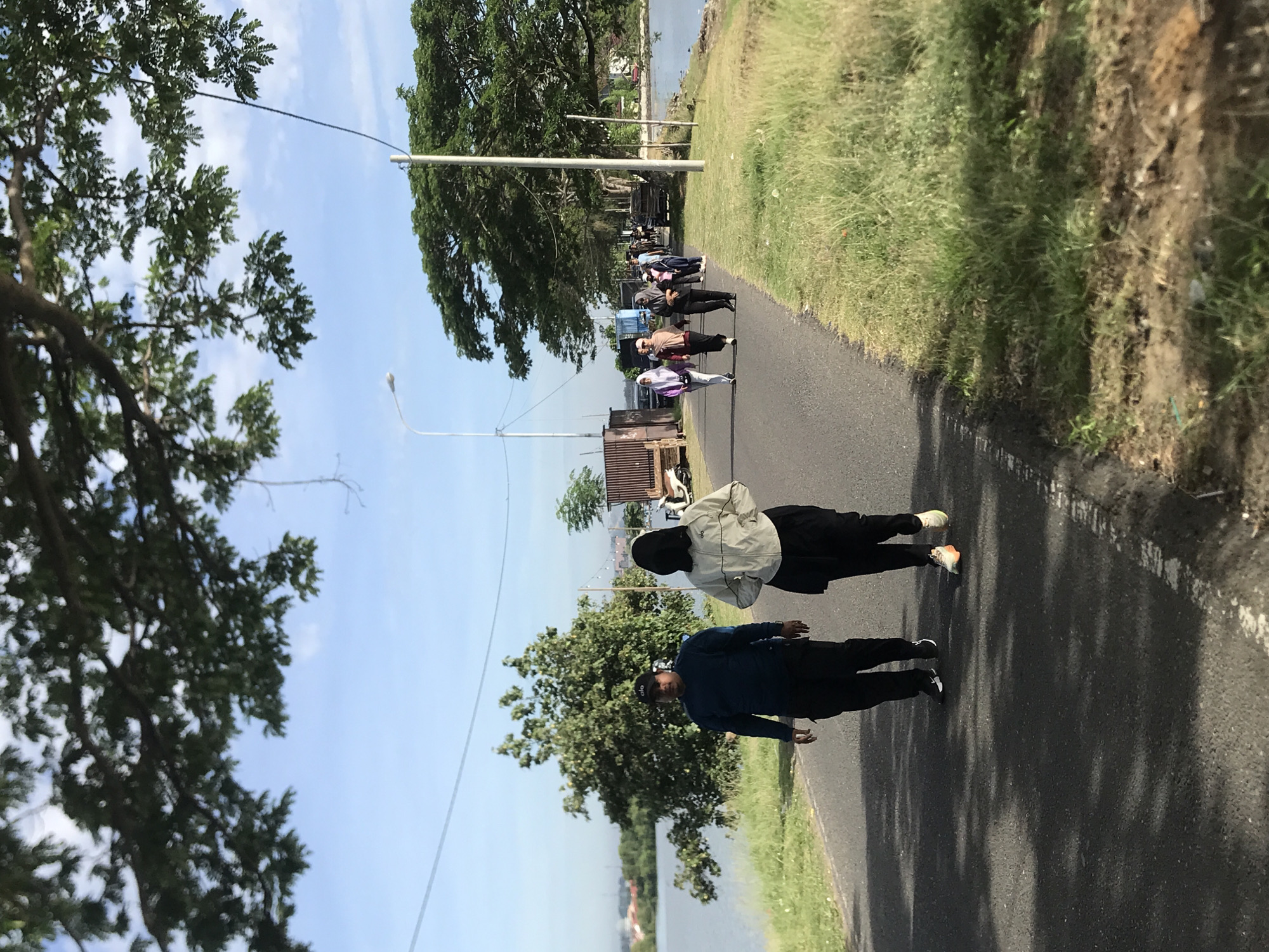 The image captures a sunny day in Lhokseumawe, Indonesia. Two people are walking along an asphalt road beside a body of water, possibly a lake or coastal area. The man in the front is wearing a blue jacket, black pants, and a dark cap. Behind him, a woman is wearing a light-colored jacket, black pants, and a hijab. They appear to be walking in the same direction.
The road is lined with green grass and trees on both sides. Further down the road, a group of people is walking. There are also some parked vehicles and structures, including what appears to be a small container, near the road. A few utility poles are visible along the roadside. The sky is blue with scattered clouds.