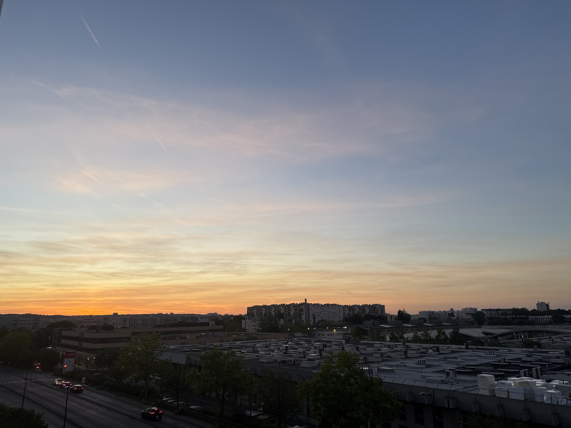 An aerial, wide-angle shot captures a city skyline at dusk. The sky transitions from a deep blue at the top to a soft pink and vibrant orange near the horizon. Wispy clouds are present, some with a light pink hue. A single contrail is visible in the upper left quadrant of the sky. Below the sky, a dense urban landscape unfolds. The middle ground is dominated by various buildings, including a large, multi-story apartment complex with many windows, and several other mid-rise structures. In the foreground, a large, flat-roofed industrial building complex is visible, with numerous ventilation units or skylights on its roof. A tree-lined street runs along the left side of the frame, with traffic signals and several cars driving on the road. The streetlights are illuminated, and the overall lighting suggests twilight conditions. The scene appears to be a view from an elevated position, possibly a building or a bridge, looking out over the city as the day ends.