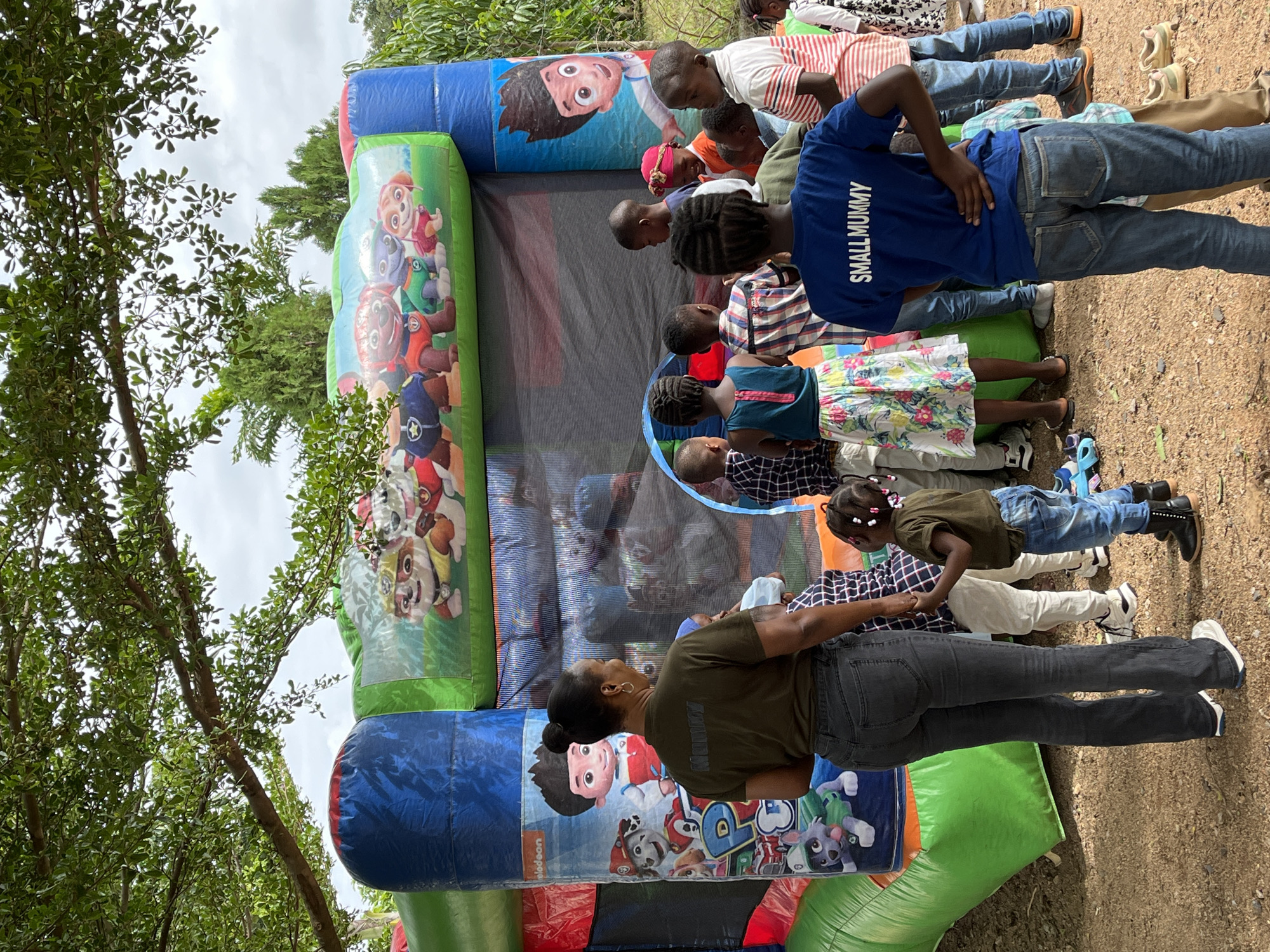 The scene is an outdoor gathering in Kuru Karama, Nigeria, featuring a multi-colored inflatable bounce house surrounded by children and adults under an overcast sky.
The central object is a large, vibrant inflatable bounce house, predominantly green and blue with red accents. The upper sections display images of characters from the "Paw Patrol" animated series, including Chase, Rubble, Skye, and Zuma, along with the Nickelodeon logo. The sides of the bounce house feature larger graphics of "Paw Patrol" characters and Ryder, with a black mesh netting enclosing the main bouncing area.
Approximately 15-20 children, ranging from toddlers to pre-teens, are gathered around the bounce house. Many are observing the entrance or waiting. A dark-skinned adult woman stands near the entrance with her back partially to the camera, wearing an olive-green t-shirt with "MUMMY" printed in lighter text on the back, dark grey jeans, and a silver hoop earring. Her hair is pulled back in a bun. She appears to be supervising the children or assisting with access to the bounce house.
In the foreground on the right, a dark-skinned boy stands with his back to the camera, wearing a blue t-shirt with "SMALL MUMMY" written in white capital letters across the back, and blue jeans. He has his hands on his hips. To his right, another dark-skinned boy wears a red and white striped polo shirt and blue jeans. A dark-skinned girl, partially visible in the foreground on the left, wears a floral skirt and a blue top. A young child, possibly a toddler, stands next to the adult woman, dressed in a dark t-shirt, blue jeans, and dark boots. Other children are dressed in various casual clothing.
The ground is composed of reddish-brown dirt or sand. Large trees with dense green foliage create an overhead canopy, providing shade. Additional trees and vegetation are visible in the background. The sky visible through the tree canopy is overcast and light grey. Footwear, including sandals and shoes, is placed on the ground near the bounce house, indicating that children have removed them before entering or are about to.