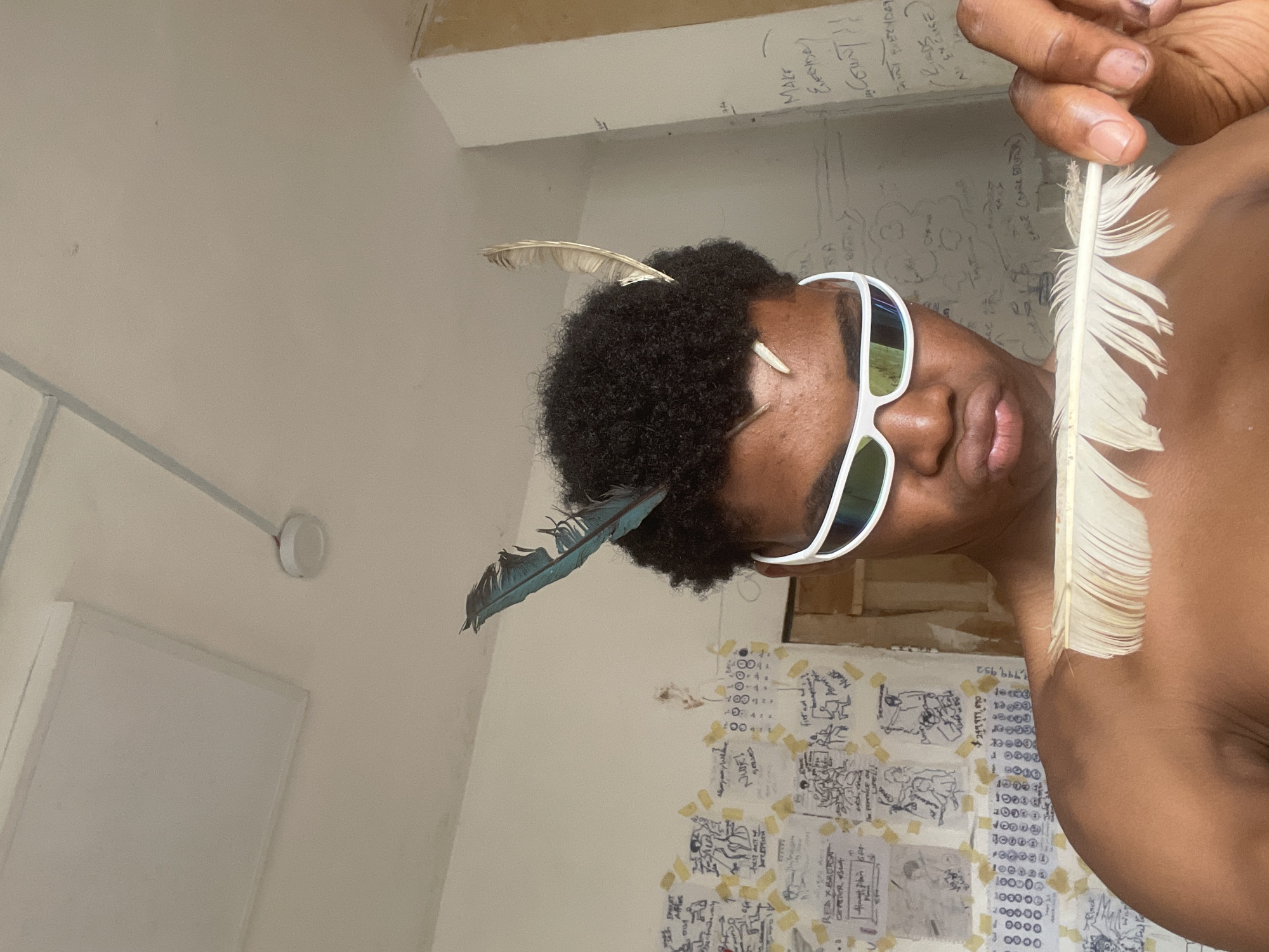 The image is a close-up, self-taken photograph of a young Black person wearing white, futuristic-style sunglasses and sporting two feathers in their curly hair, one blue and one beige. They are holding a large, white feather up to their face and chest. The background appears to be an interior space with walls covered in various papers with handwritten notes, drawings, and what look like diagrams. One wall has some text written directly on it, including "MAKE EVERYDAY COUNT!!" and "BIRDS IN CU..." and "EXCUSE ME". The ceiling is visible in the upper part of the image, with a rectangular light fixture and a smoke detector. The person's skin is dark and their shoulders are bare. The lighting is bright and seems to come from above. The context of Ado, Nigeria is not visually evident from the image itself; there are no landmarks, specific architectural styles, or other cultural cues that would place it in that specific city.