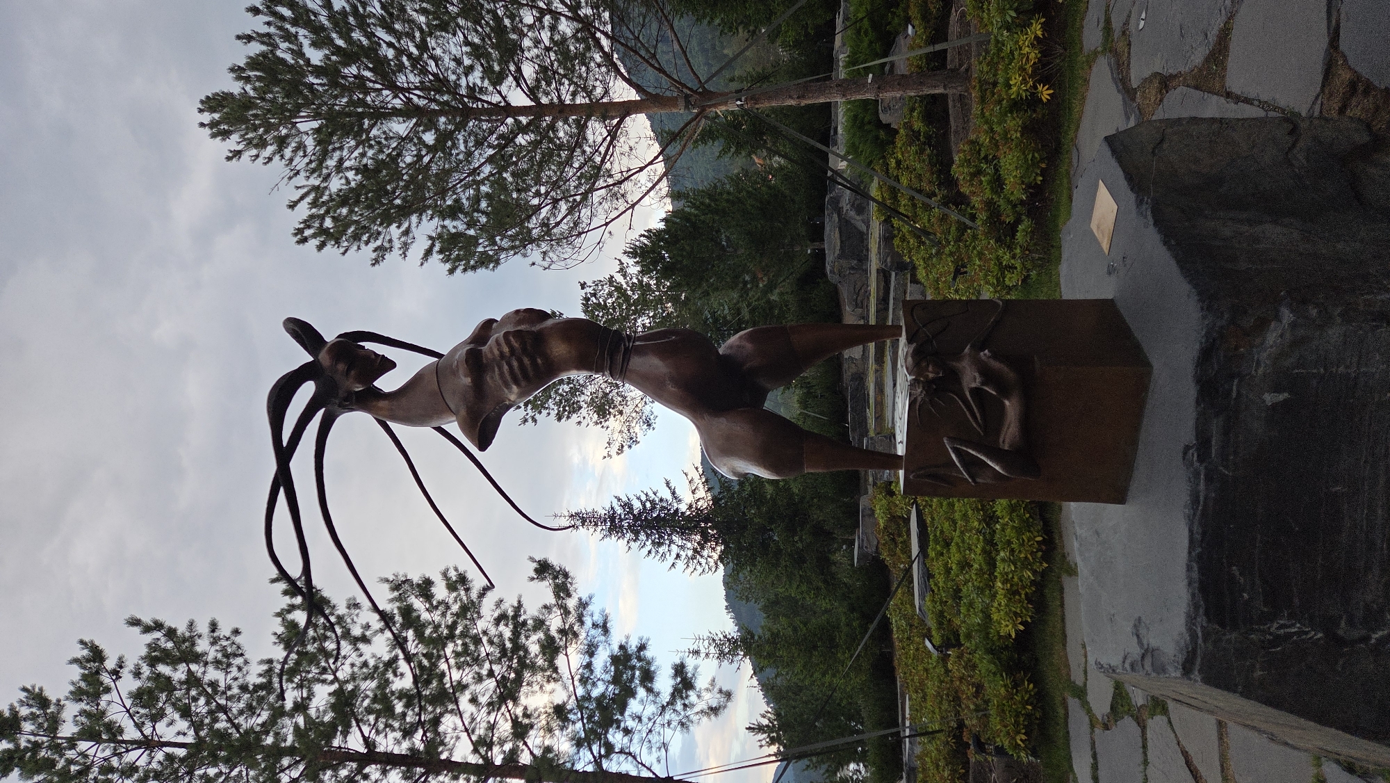 A bronze sculpture of a mythical creature stands in an outdoor park. The creature has the head and upper torso of a human-like figure with elongated, tendril-like appendages extending from its head. Its body is slender with muscular legs, and it is positioned in a dynamic, bent posture. The sculpture is mounted on a square bronze pedestal that features a relief of a reclining nude figure on one of its sides. This pedestal, in turn, rests on a large, rough-hewn dark grey stone base. The park setting includes lush green bushes, scattered rocks, and mature pine trees. In the background, a body of water can be seen on the left, with rolling hills and a cloudy sky. This sculpture is located in the city of Ozernoye (Mayskiy), Russia.