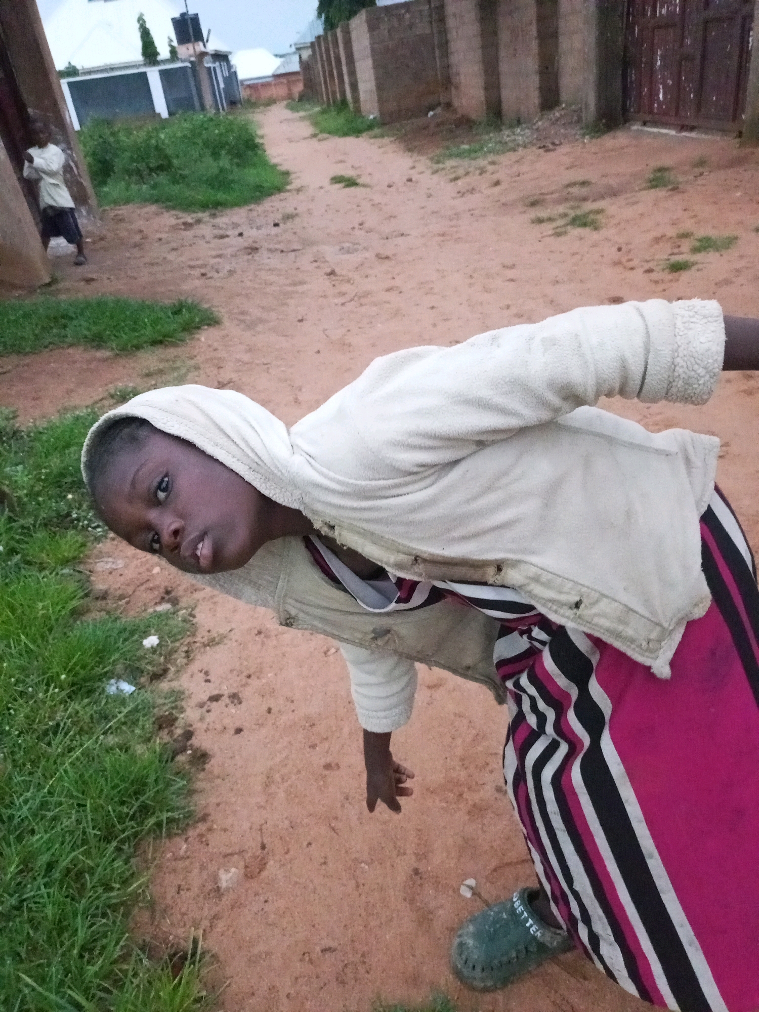 The image depicts a child wearing a white hoodie and colorful striped pants, crouching outdoors on a dirt path in what appears to be the city of Tempure, Nigeria. The child is looking towards the camera with a neutral expression. In the background, another child is visible, standing near a building. The environment is characterized by dirt roads, patchy grass, and buildings with brick and metal fences. The overall scene suggests a casual, everyday moment in a residential area.