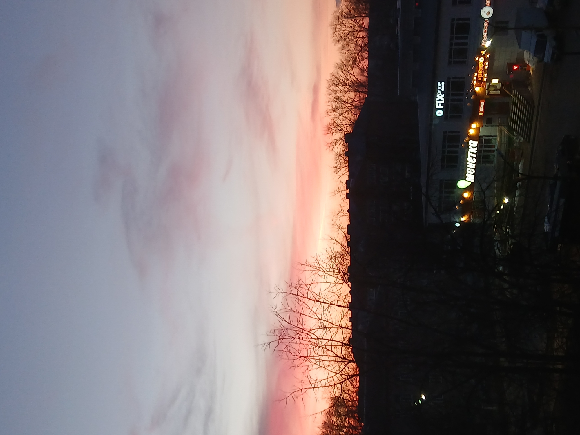 The image presents an evening scene, capturing a view over the city of Chaikovsky, Russia. The sky displays a gradient of colors, ranging from light gray to vibrant pinks and oranges, indicative of a sunset or sunrise. Silhouetted trees with bare branches are visible against the colorful sky.
On the right side of the image, a building stands, illuminated with various lighted signs in Russian. The signs read "Fix Price" and "Monetka," indicating these are the names of businesses within the building. The "Monetka" sign is particularly prominent, suggesting it is a store or shop. There is a lit crosswalk sign near the storefront.
The scene appears to be from an elevated viewpoint, offering a glimpse of the surrounding urban landscape at dusk. Cars are parked near the building. Overall, the image captures the interplay of natural light and urban features in a city setting.