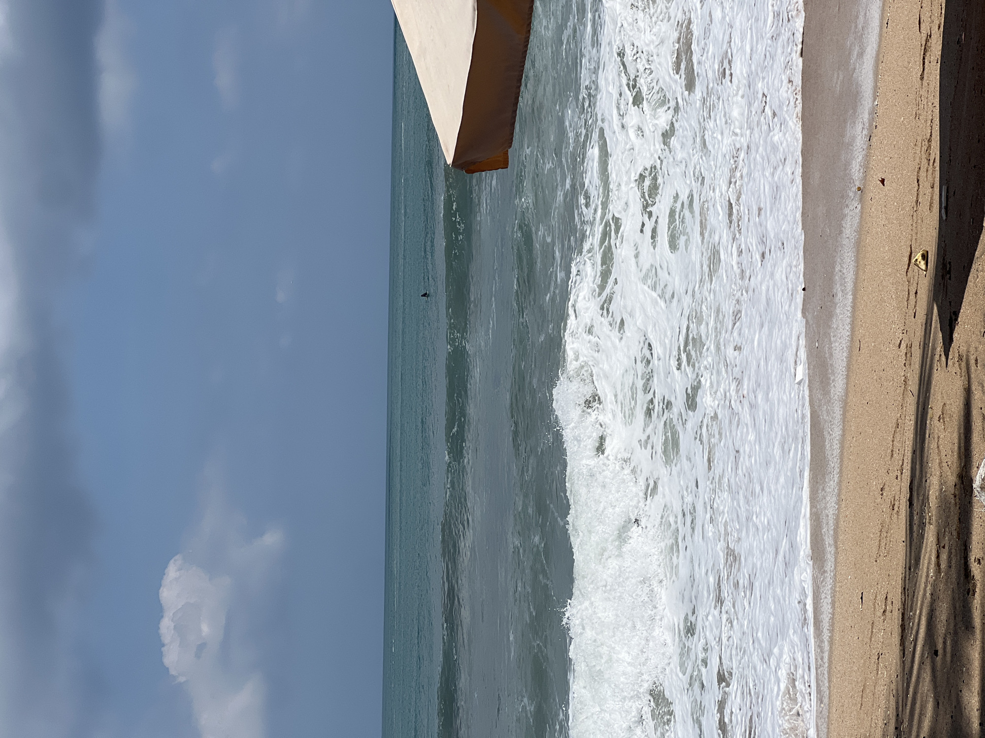 A bright, sunny day at Kuta Beach, Indonesia, is depicted in this image. The foreground features a sandy beach with shadows cast by an unseen object, possibly a beach umbrella or trees. A piece of yellow debris is visible in the sand. The ocean is active with waves breaking on the shore, creating white foam that extends onto the sand. In the distance, a lone surfer is visible in the turquoise water, and a portion of a beige beach umbrella is in the right side of the frame. The sky is partly cloudy with a mix of blue sky and gray clouds.