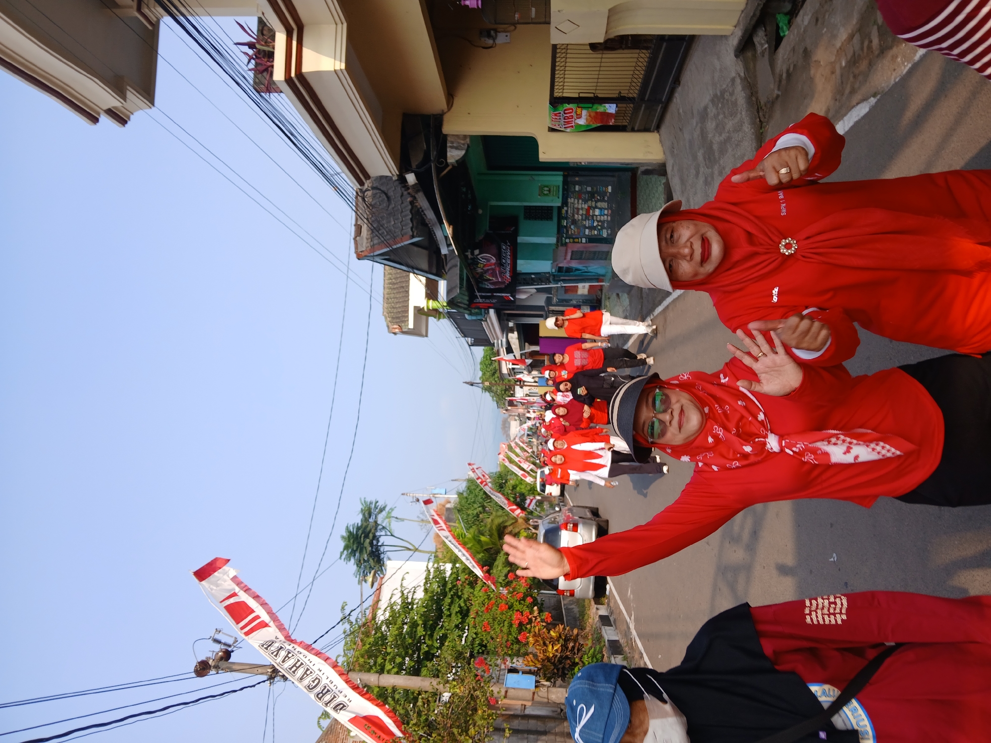 The image depicts a group of people, predominantly women, walking down a street in Ambarawa, Indonesia. The event appears to be a celebratory procession, possibly for Independence Day, as evidenced by the numerous red and white Indonesian flags and banners displayed along the street. The people are mostly dressed in red attire, with many wearing headscarves. The overall atmosphere seems festive and communal. The buildings lining the street suggest a residential or mixed-use area, with some houses and small shops visible. The sky is clear and blue, indicating it's daytime, likely morning given the quality of light. The road is paved, and there are power lines and trees present, typical of a semi-urban or urban setting.