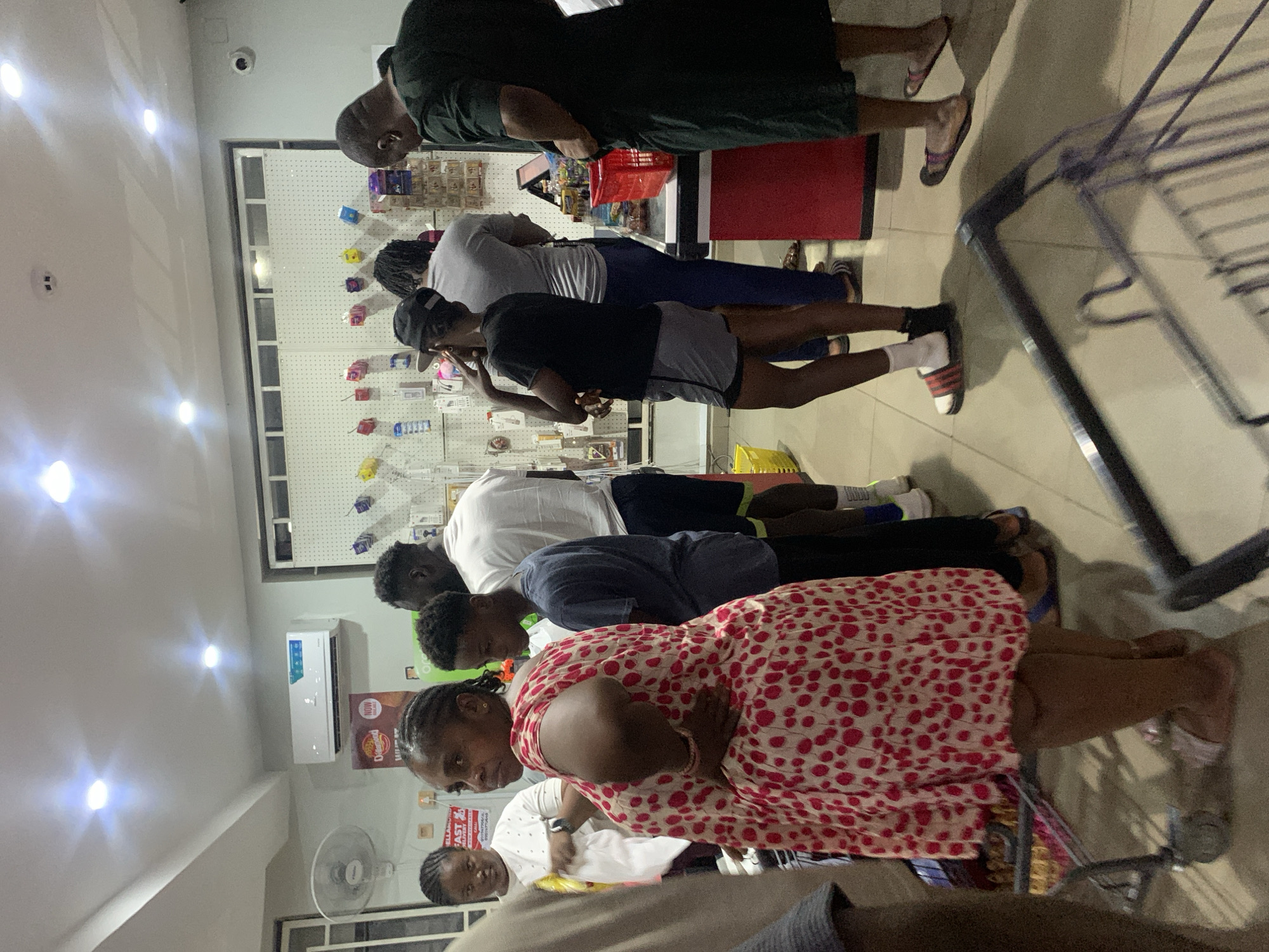 The image shows a group of people inside a store, possibly a supermarket or convenience store. The store is located in the city of Abuja, Nigeria. Multiple people are standing in a line looking at items on shelves. Several are wearing casual clothing, including t-shirts, shorts, and dresses. One person has a shopping cart. There is a display of small packaged items on the shelves in the background. An air conditioner is mounted high up on the wall. A fan is positioned low to the floor. Fluorescent lights provide illumination for the space.