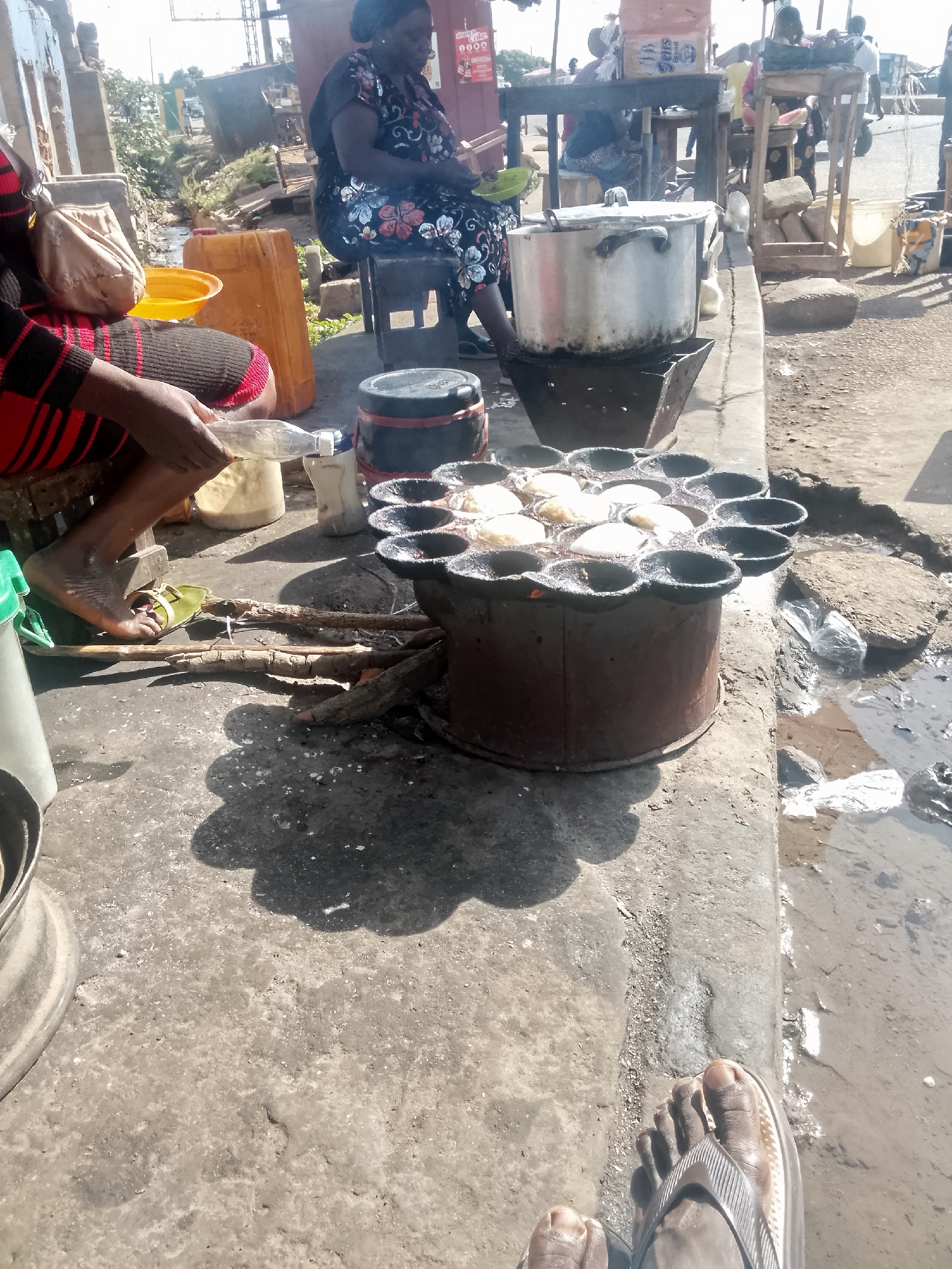 The image shows a street food scene in the city of Unknown, Unknown. A woman wearing a patterned dress sits on a stool. Next to her, another person with bare feet and wearing sandals sits preparing food. They're using a cooking contraption with multiple small circular molds, filled with what appears to be light-colored food cooking over a wood fire. There's a large metal pot on a separate stove nearby. Other people are visible in the background, sitting at tables. The surrounding area is a concrete surface, and there is a puddle of water on the side.