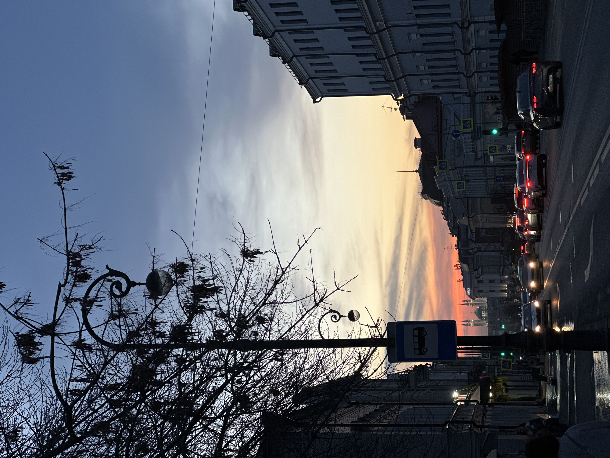 The image shows a street scene in Kazan, Russia, at dusk. The sky transitions from a dark blue-grey to a vibrant orange and yellow near the horizon. Buildings line both sides of the street, with classical architectural details. Cars are stopped in traffic on the right side of the street, their headlights and taillights visible. Streetlights are illuminated. A bus stop sign is visible on the right. In the foreground, a bare tree and an ornate lamppost create a silhouette against the sky. The street surface appears wet, reflecting the lights. The scene is generally calm and urban.