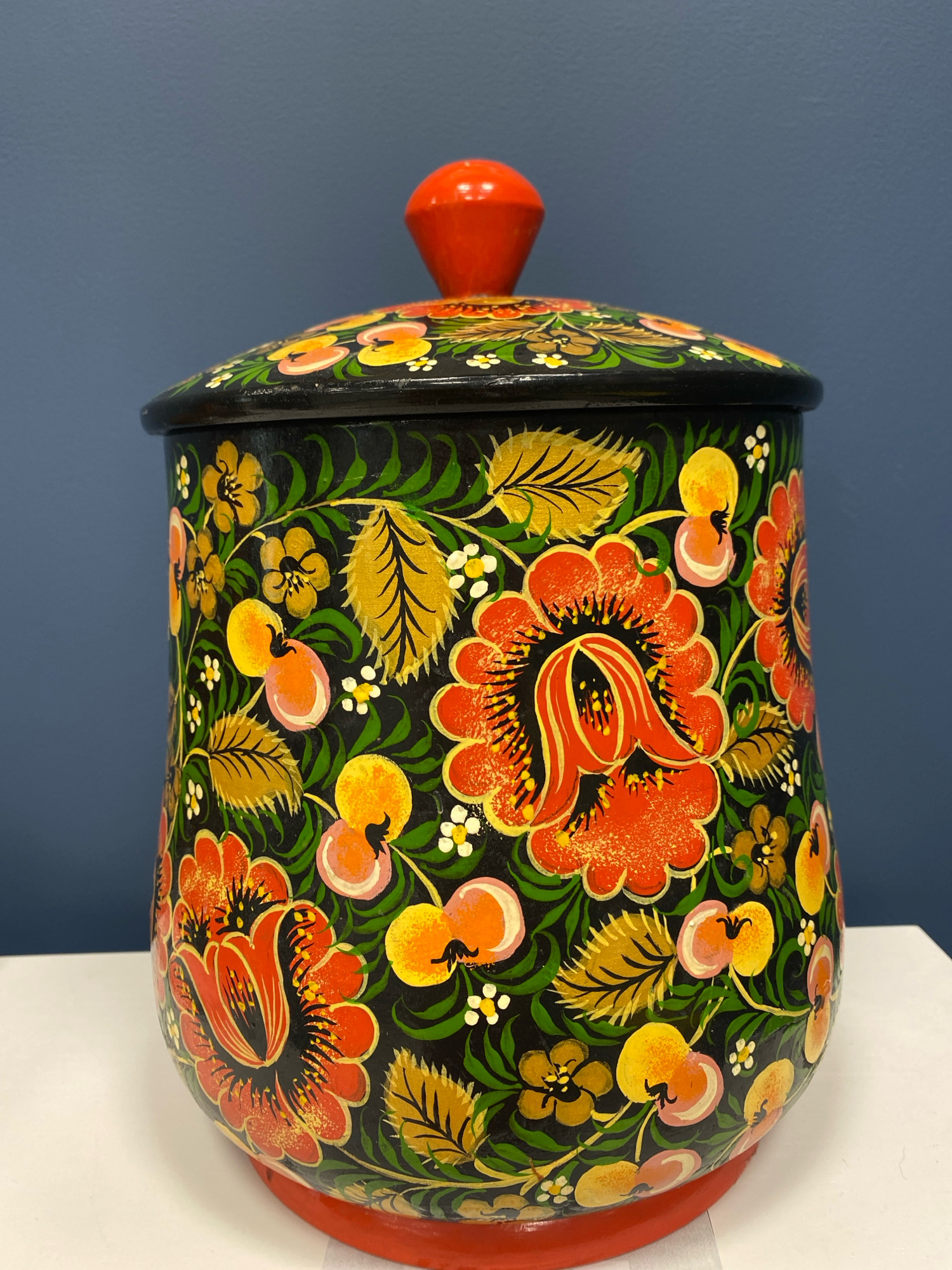 A brightly painted wooden canister with a red lid sits on a white surface in front of a dark blue wall. The canister is decorated with a black background and features a vibrant floral pattern in red, orange, yellow, and green. The flowers are stylized and detailed, with intricate petal designs. The lid has a red knob and is also adorned with a similar floral motif. The lighting is even, and the image is sharp, providing a clear view of the object. The scene appears to be indoors. The image does not contain any information about the city of Ivanteyevka, Russia.