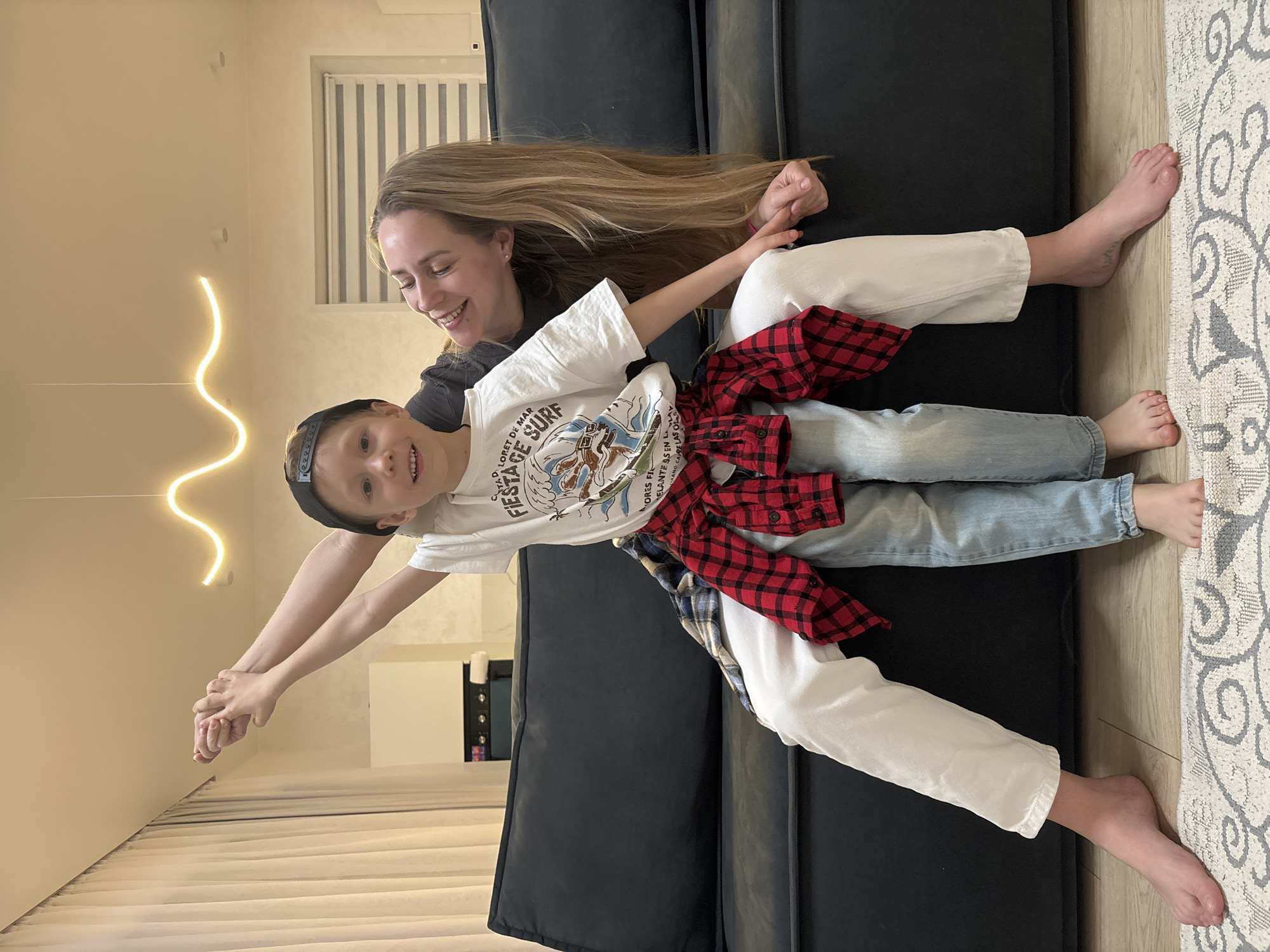 A joyful indoor scene in Chaikovsky, Russia, features a woman and a young boy playing on a dark grey sectional couch. The image is oriented with the subjects lying horizontally, holding hands and smiling brightly at the camera, conveying a sense of playfulness and happiness.
The woman, with long blonde hair cascading down her back, wears a dark t-shirt and white pants. She is barefoot. The boy, likely her son, wears a white t-shirt with a graphic design, light blue jeans, and a red and black plaid shirt tied around his waist. He has a black baseball cap worn backward and is also barefoot.
The setting appears to be a modern living room. To the left, a light-colored wall features a distinctive wavy, warm-toned LED strip light fixture. A window with white horizontal blinds is visible in the upper background. The flooring consists of light-colored wood next to a patterned rug with grey and white motifs.
Text visible on the boy's white t-shirt includes phrases such as "LORET DE MAR", "FIESTA SURF", "MORES FIL", "ELANTES EN LA", and "SANO CARELAS OL". The overall atmosphere is lighthearted and cheerful, capturing a moment of bonding between the two subjects.