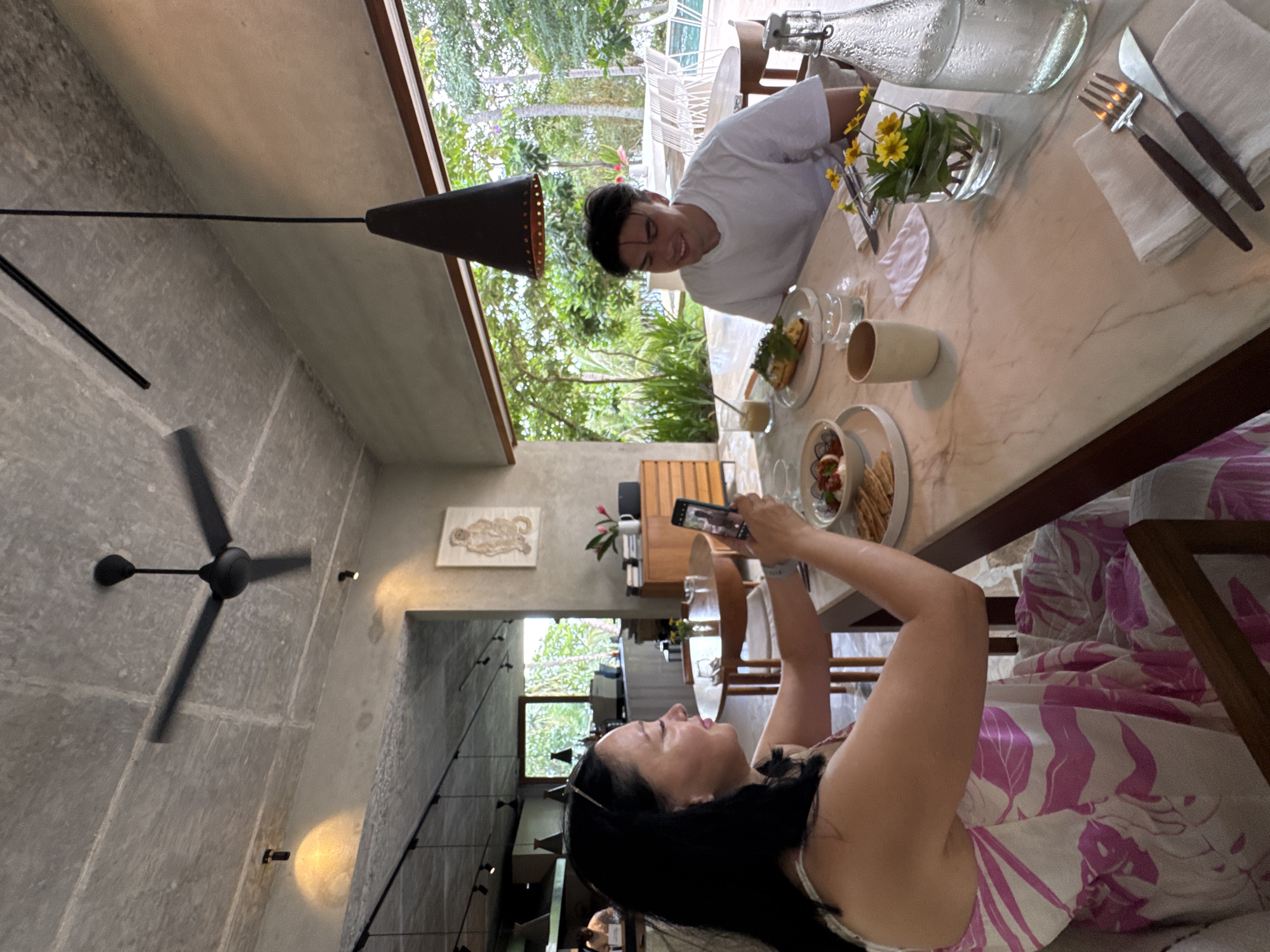 The image shows a woman and a man seated at a marble-topped table in an open-air dining area. The woman, on the left, has dark hair and is wearing a pink and white floral dress. She is holding a smartphone and appears to be taking a picture of her meal. The man, on the right, has dark hair and is wearing a white t-shirt. He is smiling and looking towards the woman. On the table are plates of food, glasses, cutlery, a vase with yellow flowers, and a bottle of water. The environment suggests a tropical setting, with lush greenery visible through the open side of the dining area, and outdoor furniture and a swimming pool in the background. The architecture features exposed concrete ceilings and large windows, common in contemporary design. The specific location is identified as Kathaluwa, Sri Lanka, suggesting a resort or restaurant in that area.